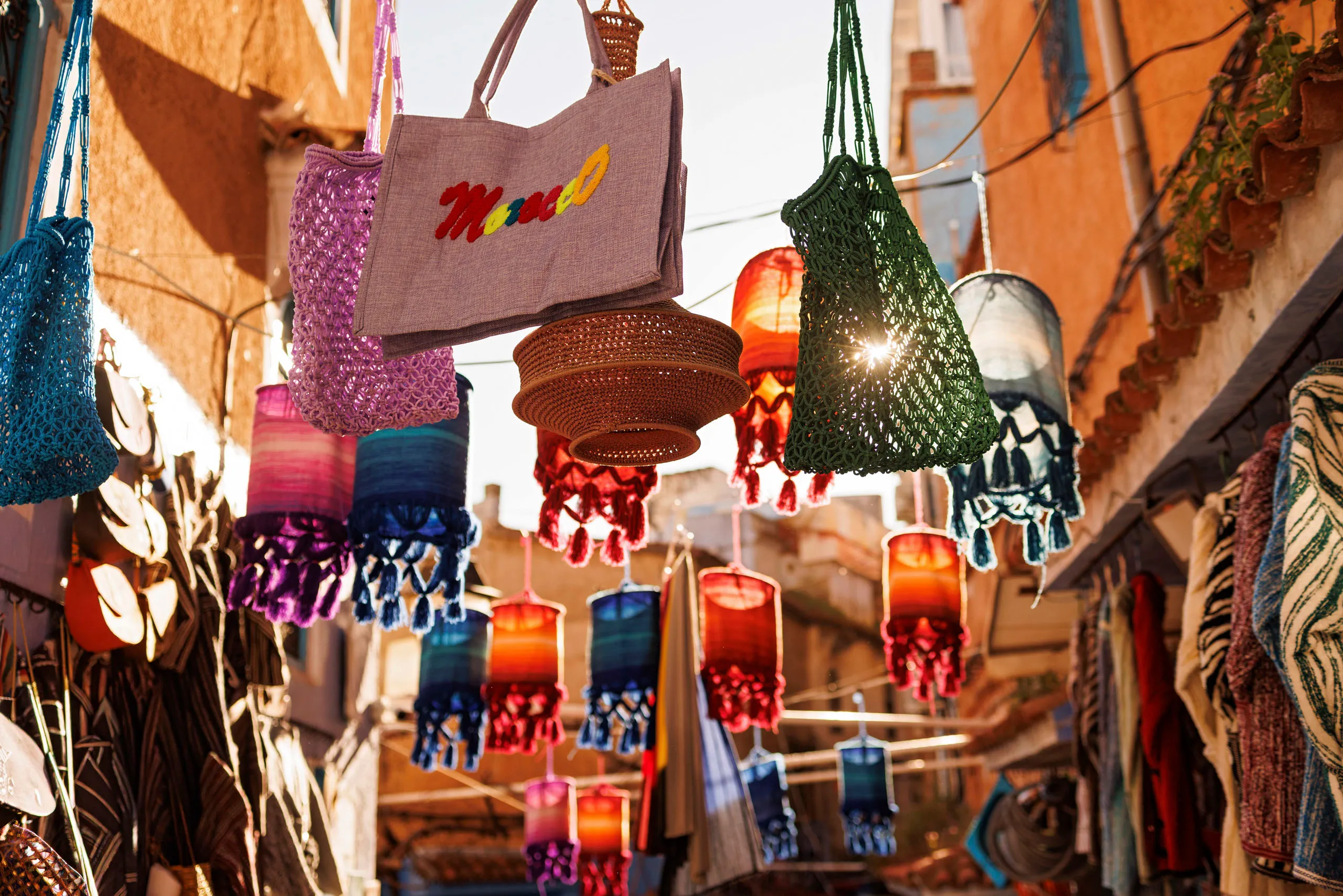 Zwischen zwei Häuserwänden einer engen Gasse hängen Schnüren, an den viele verschiedene Taschen aufgehängt sind. Dei Taschen leuchten in diversen Farben, da sie von hinten durch die Sonnen angestrahlt werden. Der Blick der Kamera ist nach oben gerichtet.