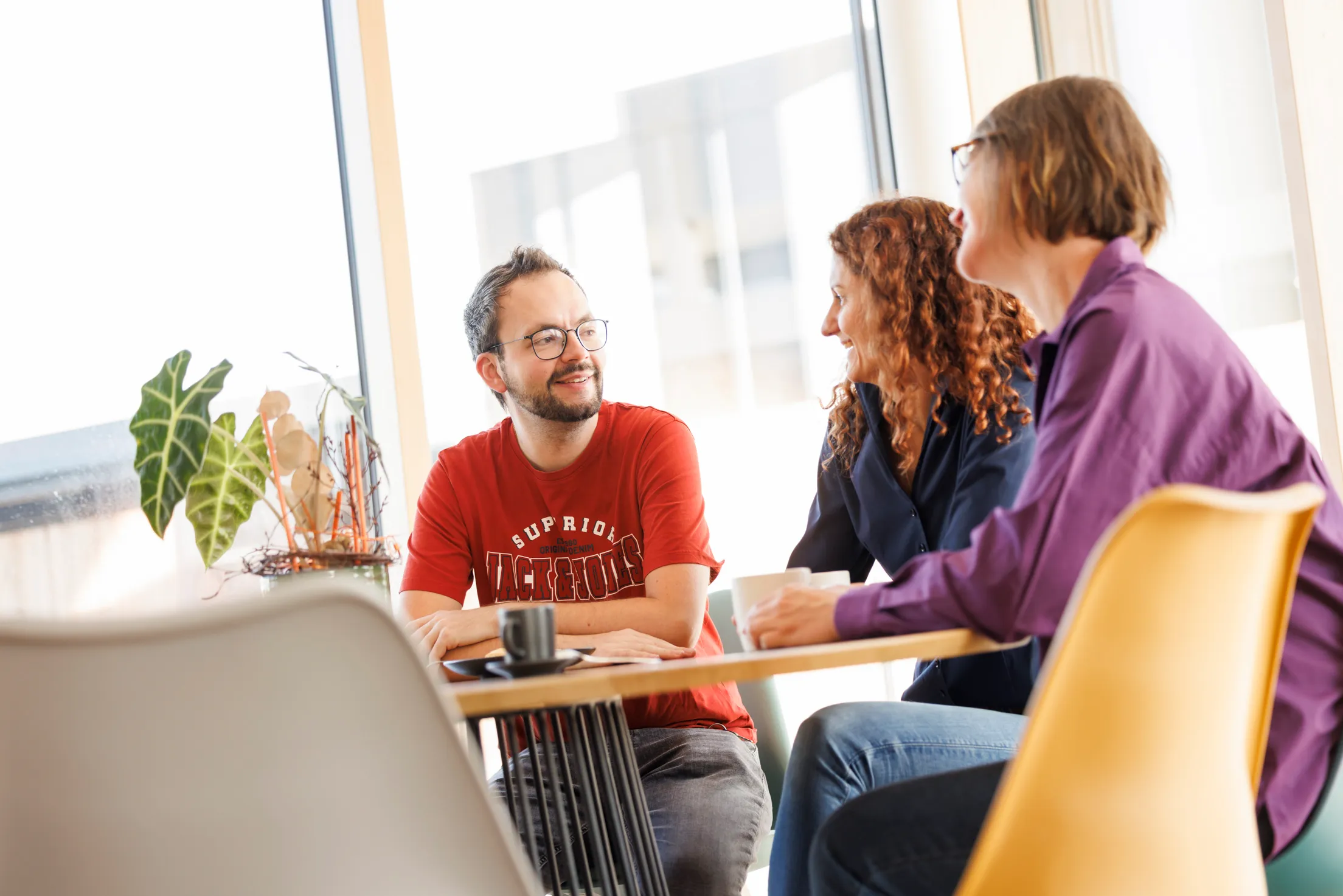 Drei Personen sitzen an einem Tisch vor einem Fenster. Sie tragen bunte Kleidung und unterhalten sich. Nur ein Mann mit Brille ist links im Bild scharf abgebildet, die beide Damen sind rechts in der Unschärfe.