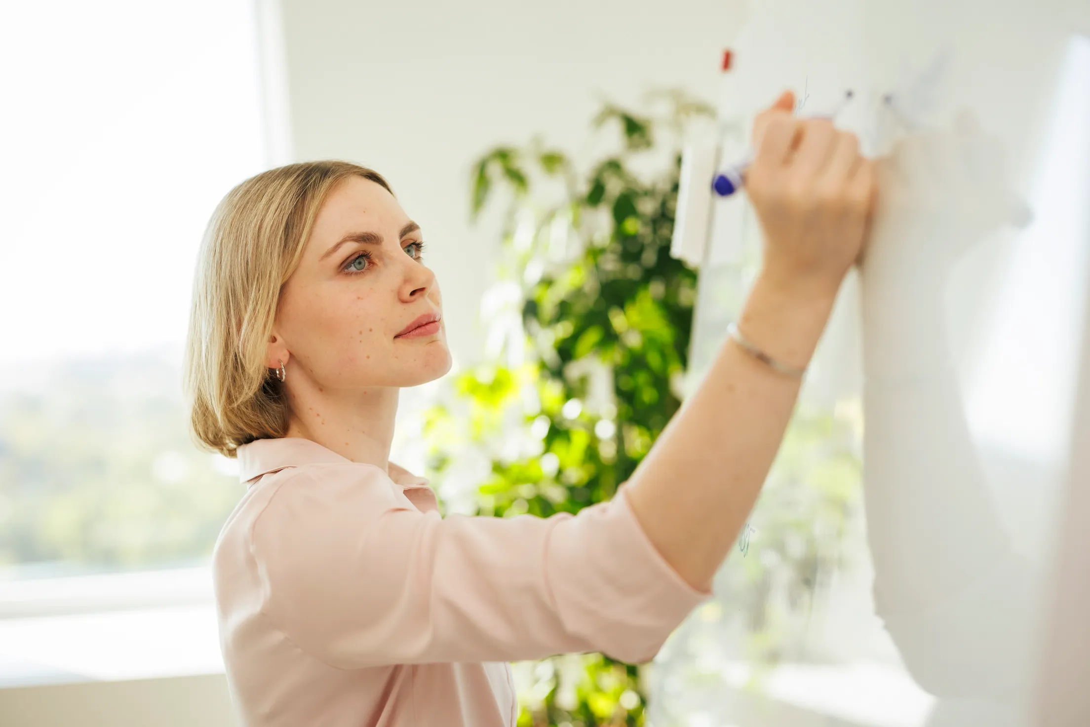 Eine blonde junge Dame ist von der Seite zu erkennen und schreibt etwas auf ein Flipchart. Im Hintergrund eine Grünpflanze