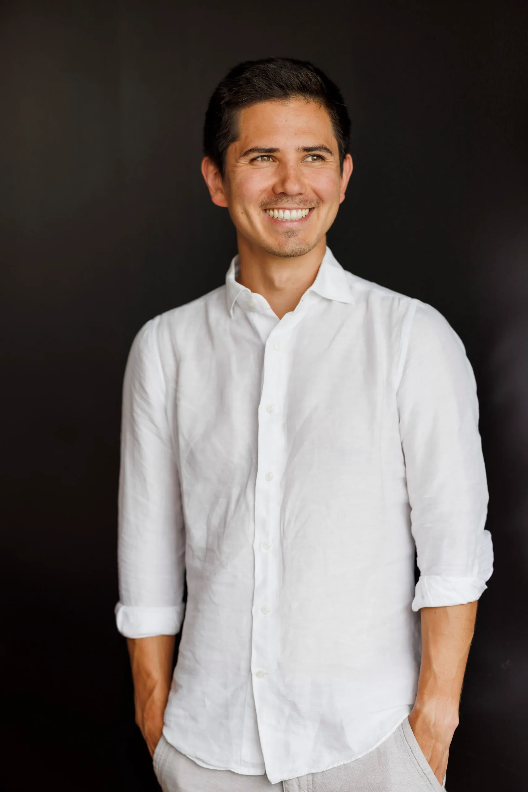 Ein junger Mann mit schwarzem Haar und weißem Hemd steht vor einem schwarzen Hintergrund. Er schaut lächelnd zu seiner linken Seite aus dem Bild.