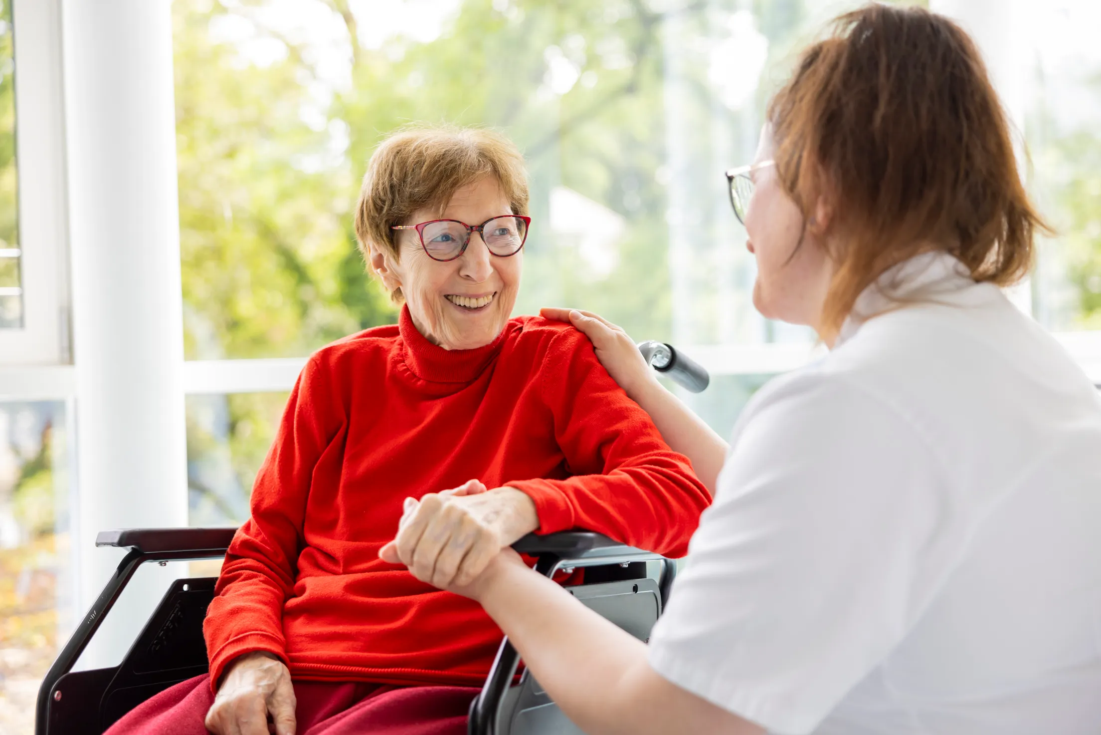 Eine Patientin mit knalligen roten Rollkragenpullover sitzt in einem Rollstuhl. Sie lächelt eine Krankenschwester an, diese hält die Hand der Dame.