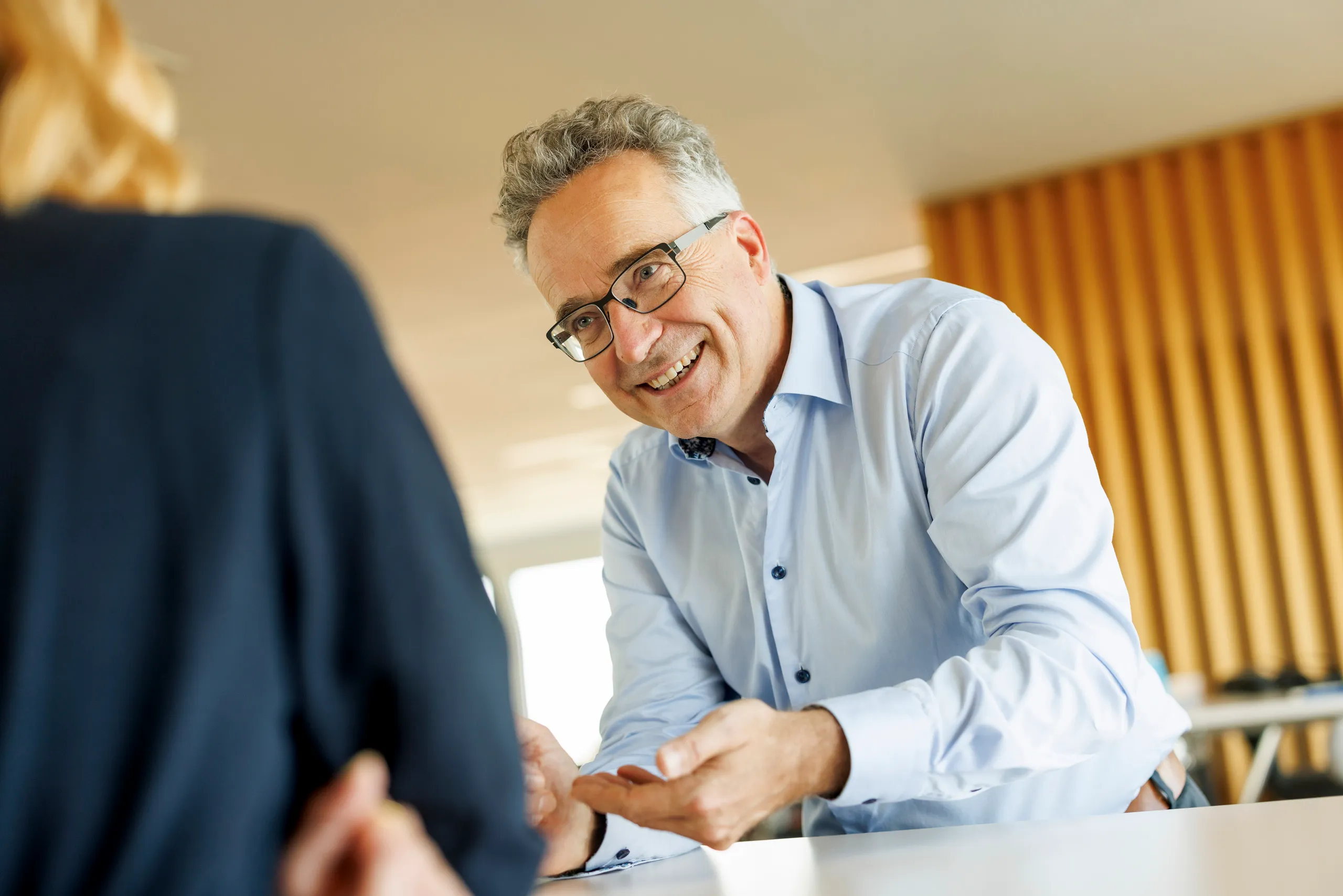 Ein Business Mann mit grauem, lockigem Haar und hellblauem Hemd ist an einem Tisch im Gespräch mit einer Person, die im Anschnitt zu sehen ist