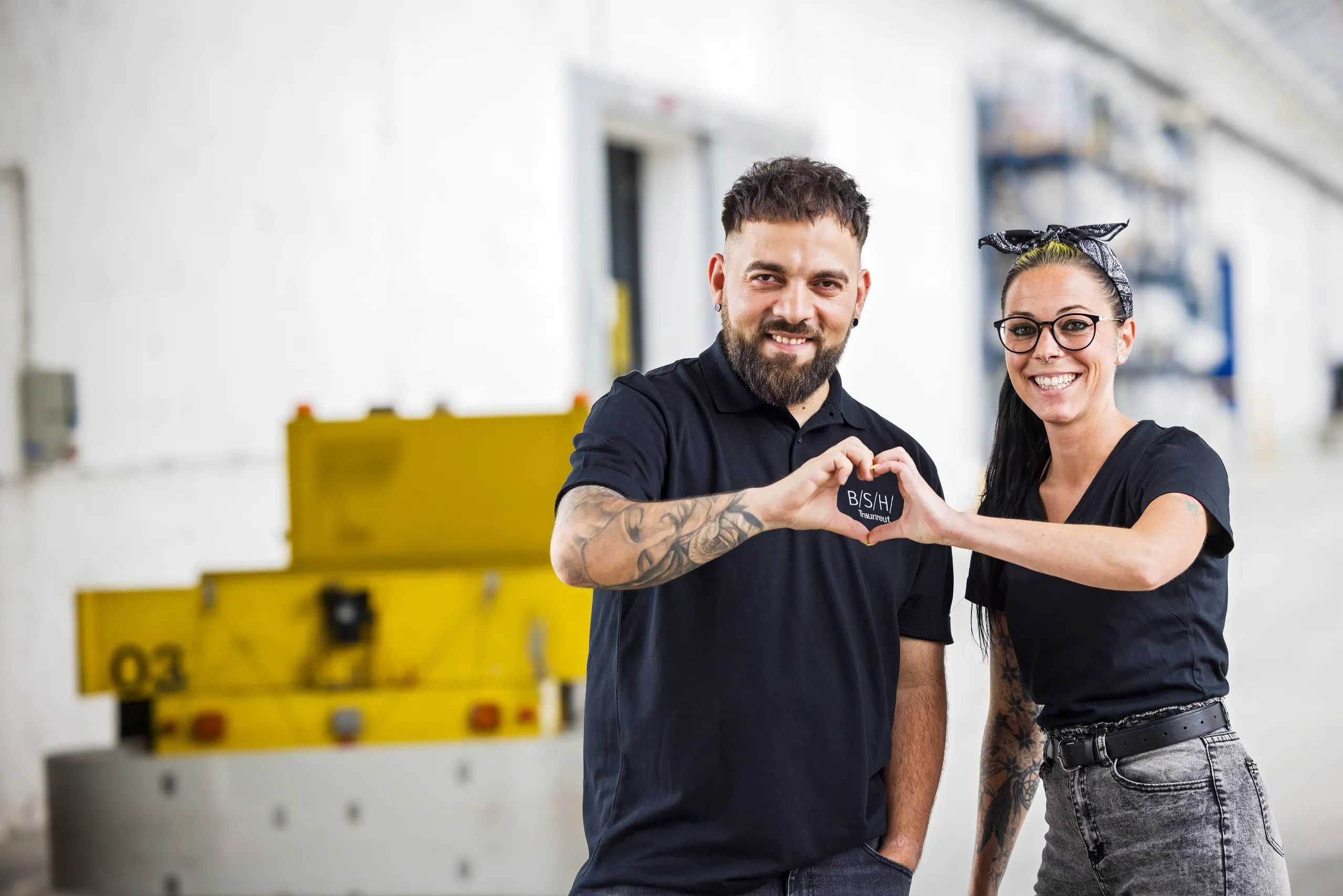Zwei Mitarbeiter von BSH stehen mit einem schwarzem Poloshirt in der Logistikhalle. Sie halten ihre Hände zu einem Herz zusammen, in der Mitte ist das BSH Logo zu sehen. Im Hintergrund ist unscharf ein gelber Roboter zu sehen.