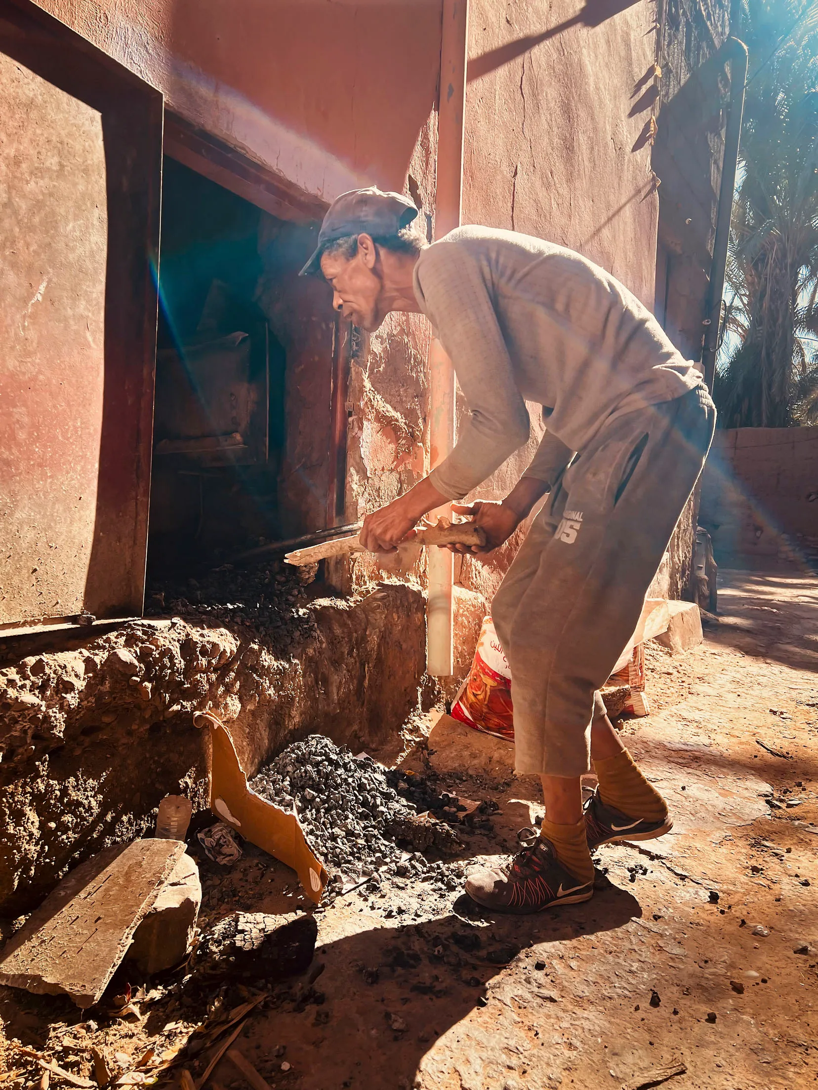 In einer Seitengasse in Zagora wirft ein Arbeiter Holz in einen Ofen der zu einer Bäckerei gehört. Er steht leicht gebeugt und trägt beige Kleidung. Die Sonne strahlt in die Kameralinse.