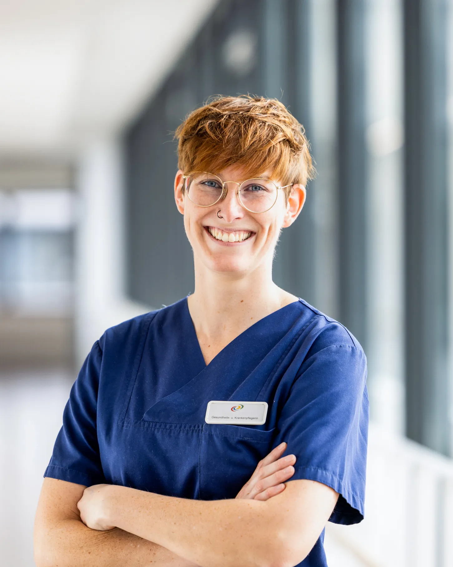Eine jüngere, sehr sympathische Krankenschwester trägt einen dunkelblauen Kasack und hat ihre Arme verschränkt. Sie schaut mit einem intensiven Lachen in die Kamera, hat kupferrotes, kurzes Haar, eine auffällige goldene Brille und im rechten Nasenloch einen Ring. Sie steht in einem hellen Klinikgang mit dunkelgrauen, großen Fenstern, welcher unscharf fotografiert ist.