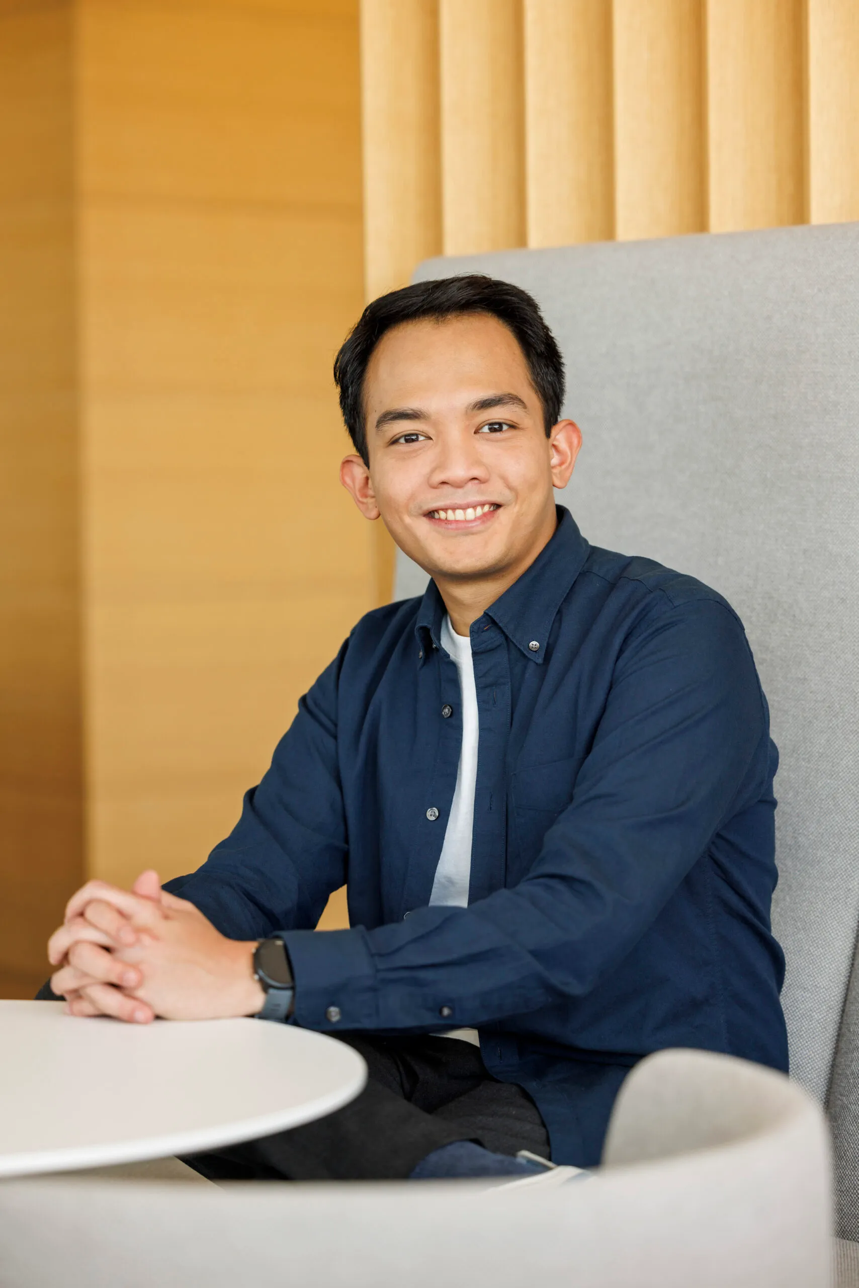 Ein junger, asiatische Mann sitzt an einem Tisch mit hellgrauer Lehne im Rücken. Er hat die Hände verschränkt und schaut sympathisch in die Kamera. Er trägt ein weißes Shirt und ein dunkelblaues Hemd