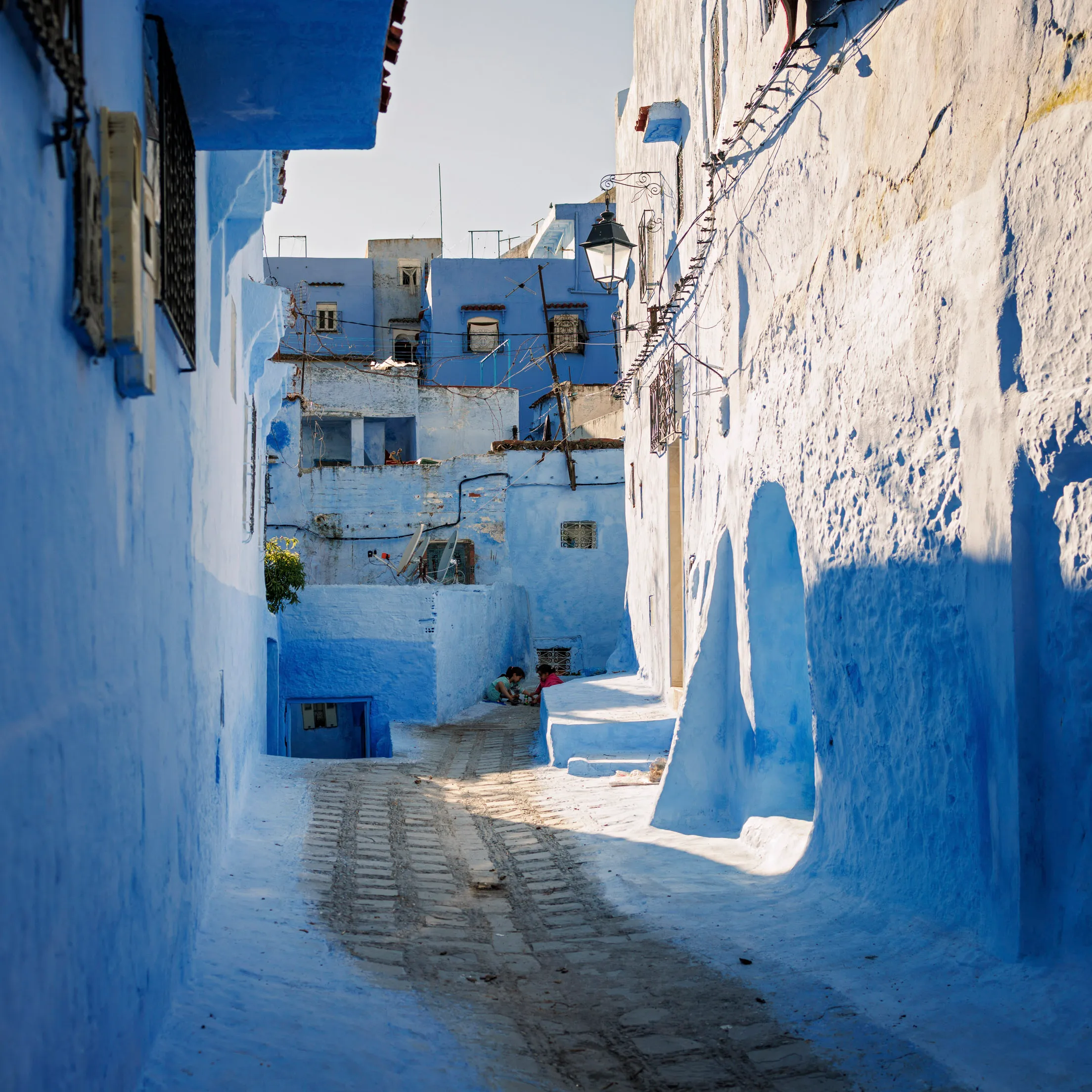 Eine Seitengasse in Chefchaouen, der blauen Stadt in Nordmarokko. Die Häuserwände sind blau angemalt und die Sonne scheint in die Gasse.