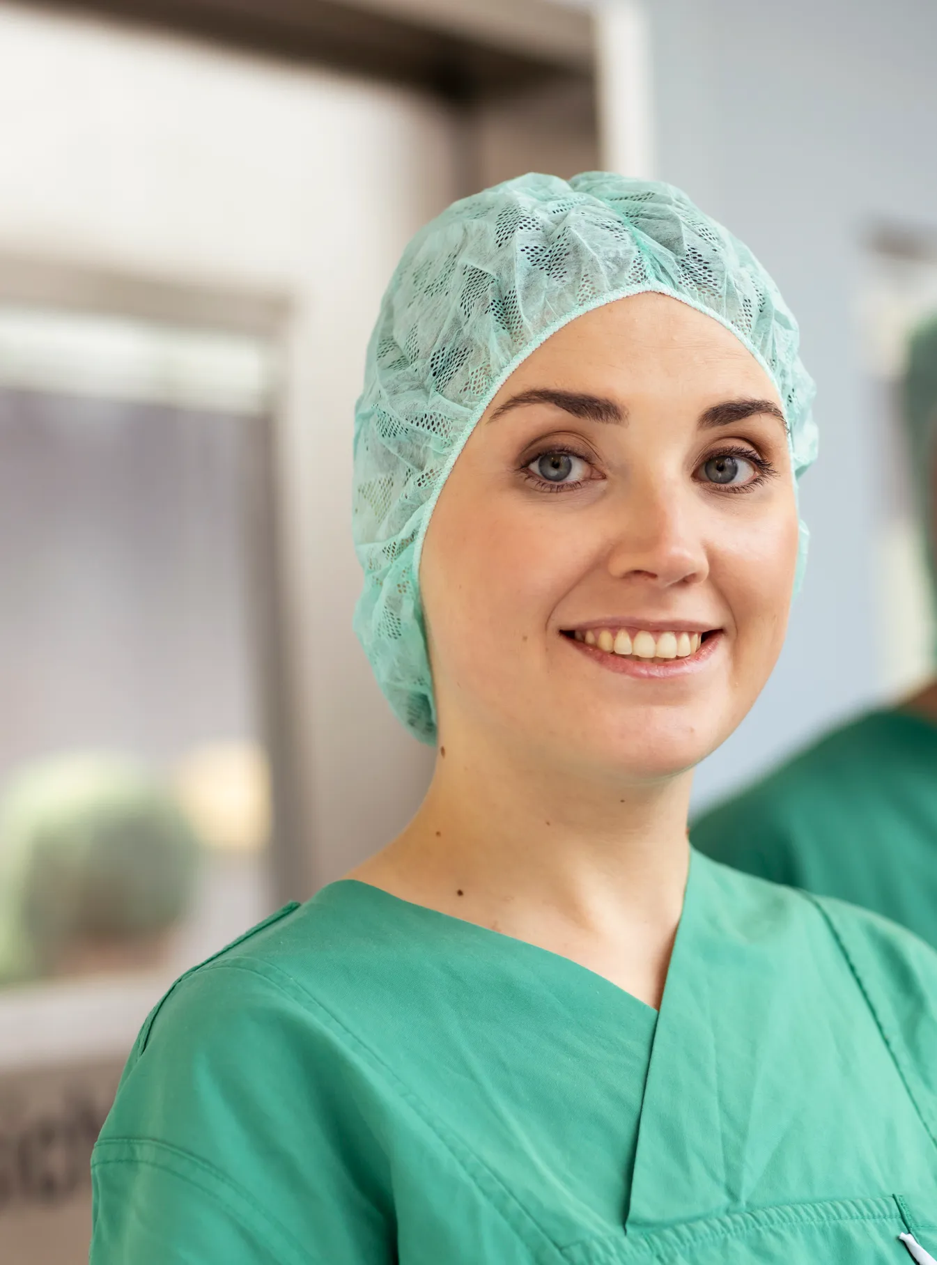 Portraits einer OP Schwester mit grüner OP Kleidung und Haube. Im Hintergrund eine silberne OP Tür.
