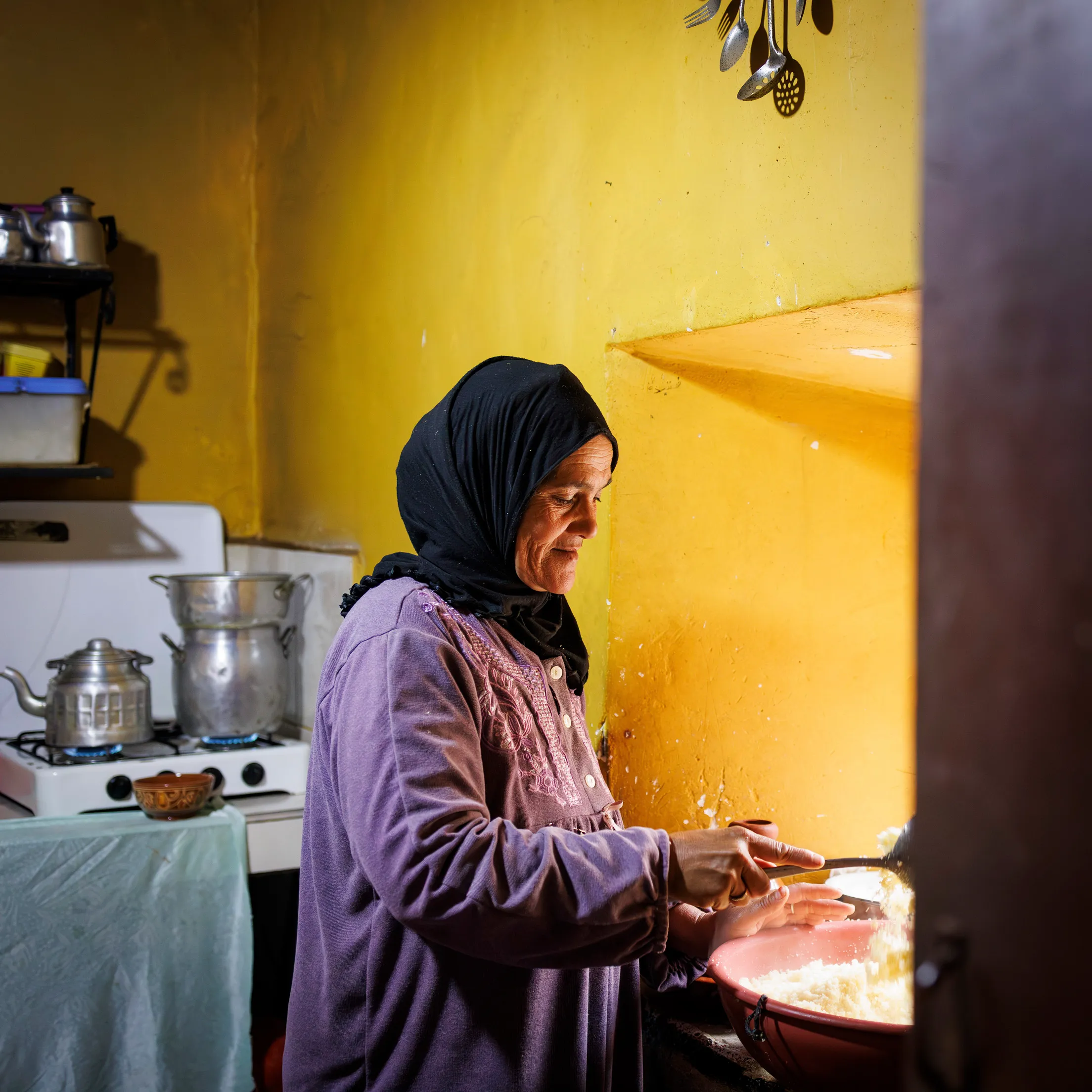 Eine marokkanische Frau mit schwarzem Hijab und lila Gewand steht in einer einfachen, gelben Küche. Sie kocht Couscous in einem großen Topf. Links hinten sind zwei Aluminiumtöpfe auf einer Kochplatte zu sehen.