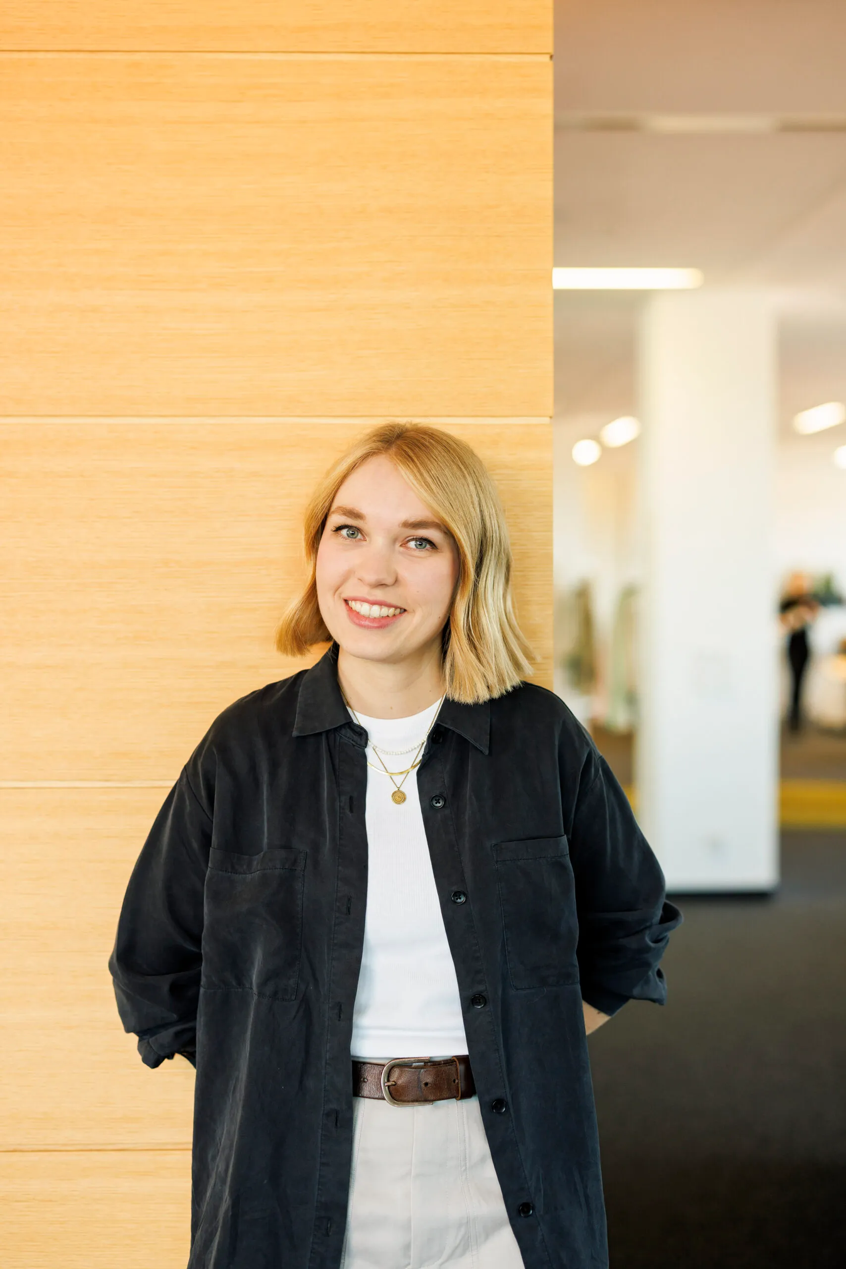 Eine junge, blonde Frau mit halblangem Haar steht mit einem schwarzen Cordhemd und die Arme nach hinten genommen, an einer hellen Holzwand. Im Hintergrund rechts ist unscharf ein Büro zu sehen