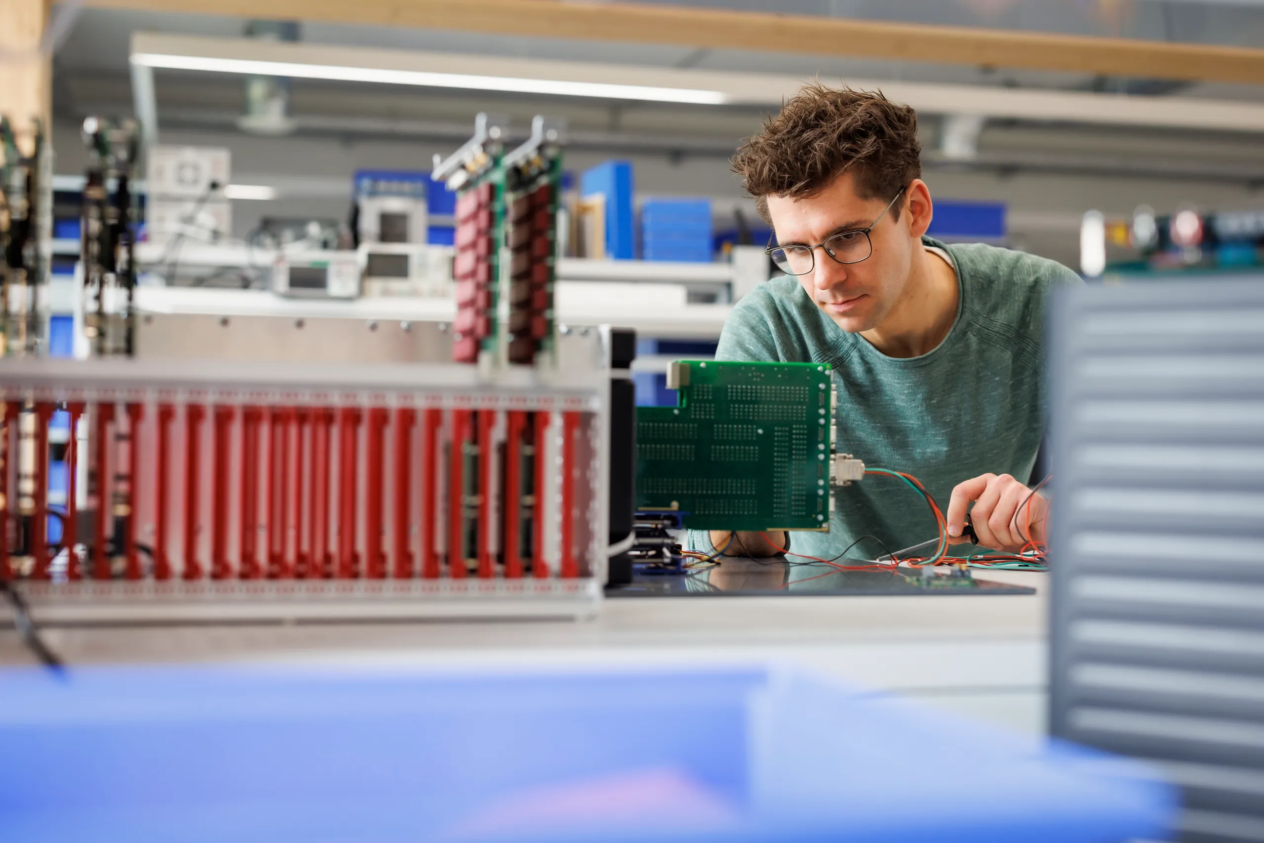 Ein Mann mit grünem Sweatshirt schaut konzentriert auf ein Halbleitersystem. Das ist ohne Verdeckung und wird gerade bearbeitet. Es ist viel blaue Unschärfe unten im Bild zu sehen.
