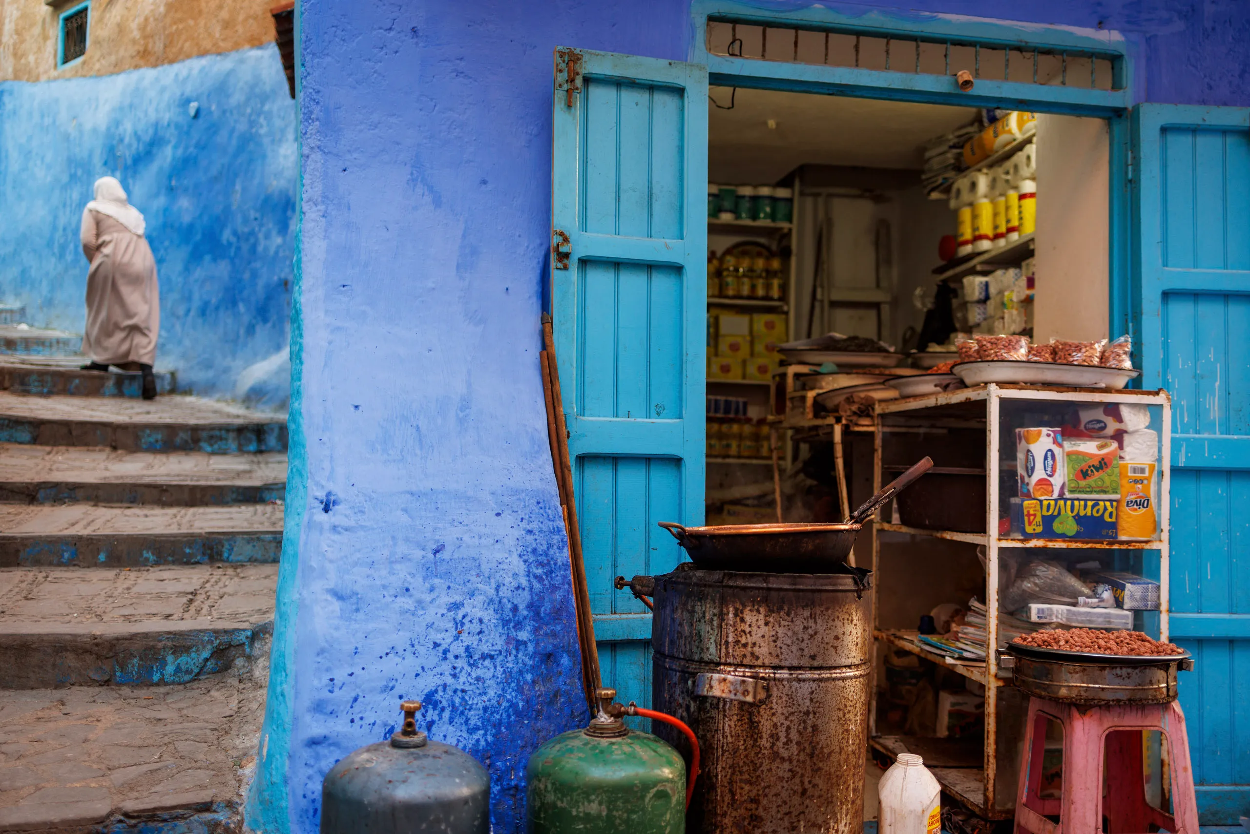 Ein Geschäft in der blauen Stadt Chefchaouen mit blauen Toren und ein paar Lebensmitteln zum Verkauf. Links davon gibt es eine Treppe, auf der eine marokkanische Frau gerade hinauf geht.