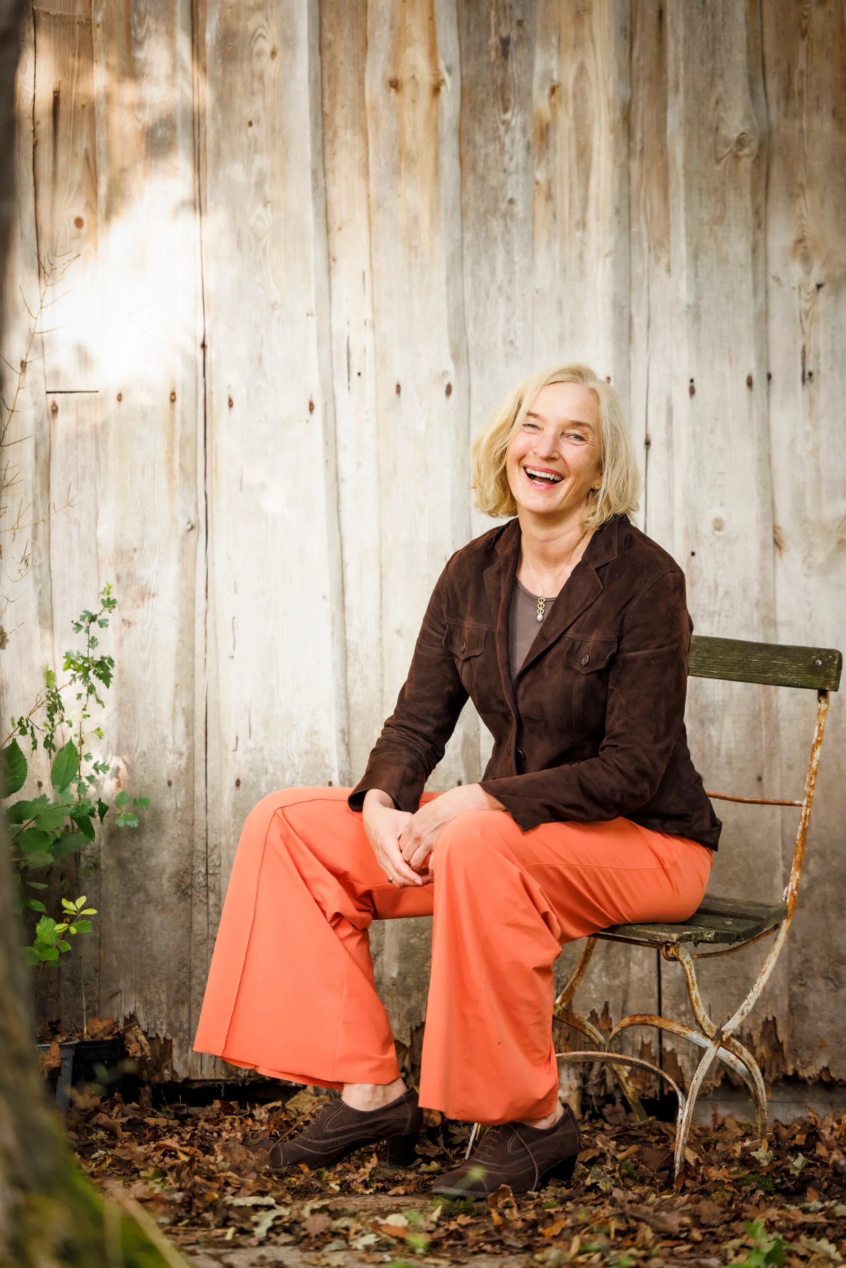 Eine Frau sitzt lachend auf einem Gartenstuhl vor einer Holzwand. Sie trägt eine knallige orangefarbene Hose und ein braunes Cordjacket.