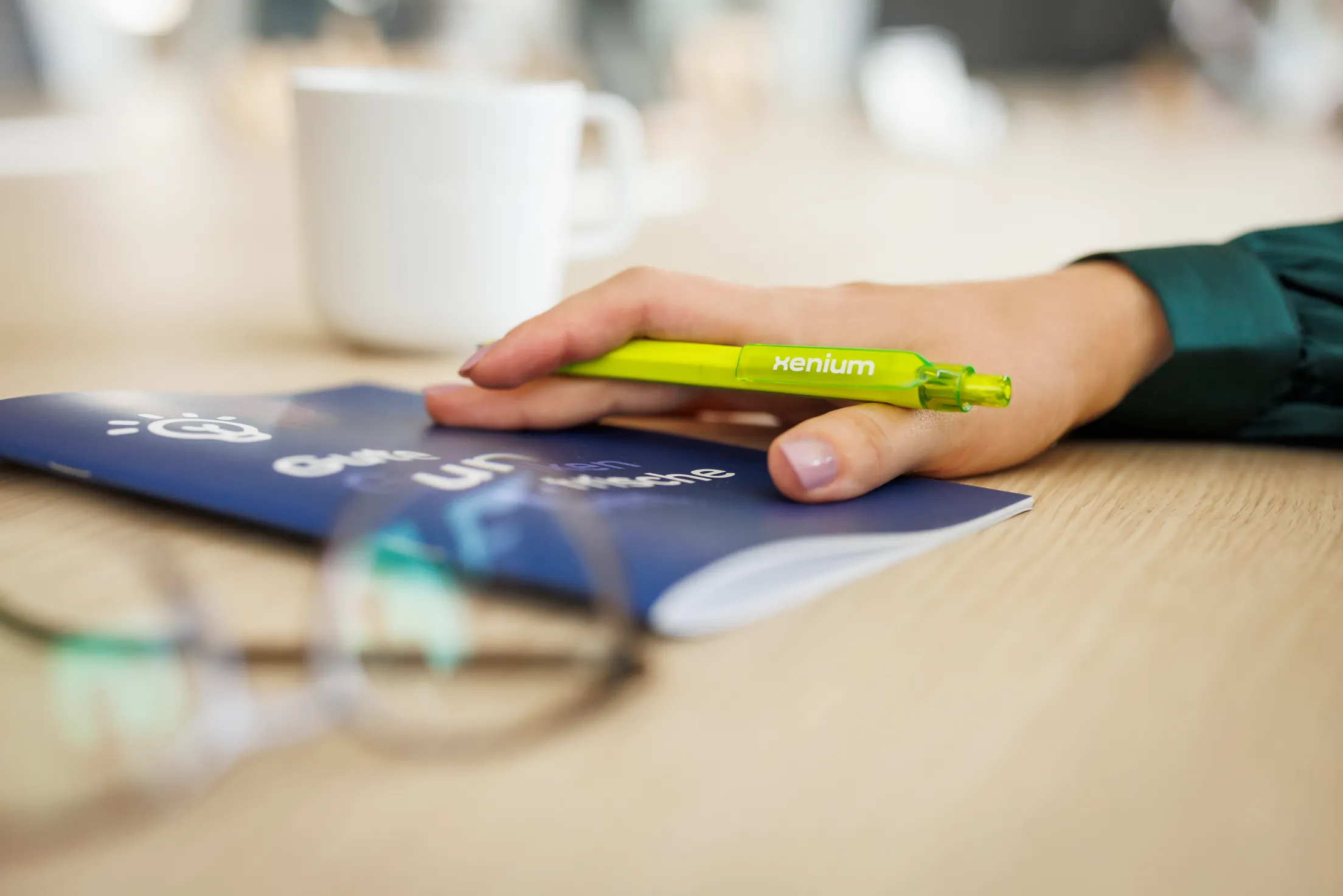 Heller Schreibtisch, ein Stillife: eine Hand mit grünem Stift, da steht Xenium darauf, liegt auf einem blauen Heft, unscharf davor eine Lesebrille, im Hintergrund eine weiße Tasse
