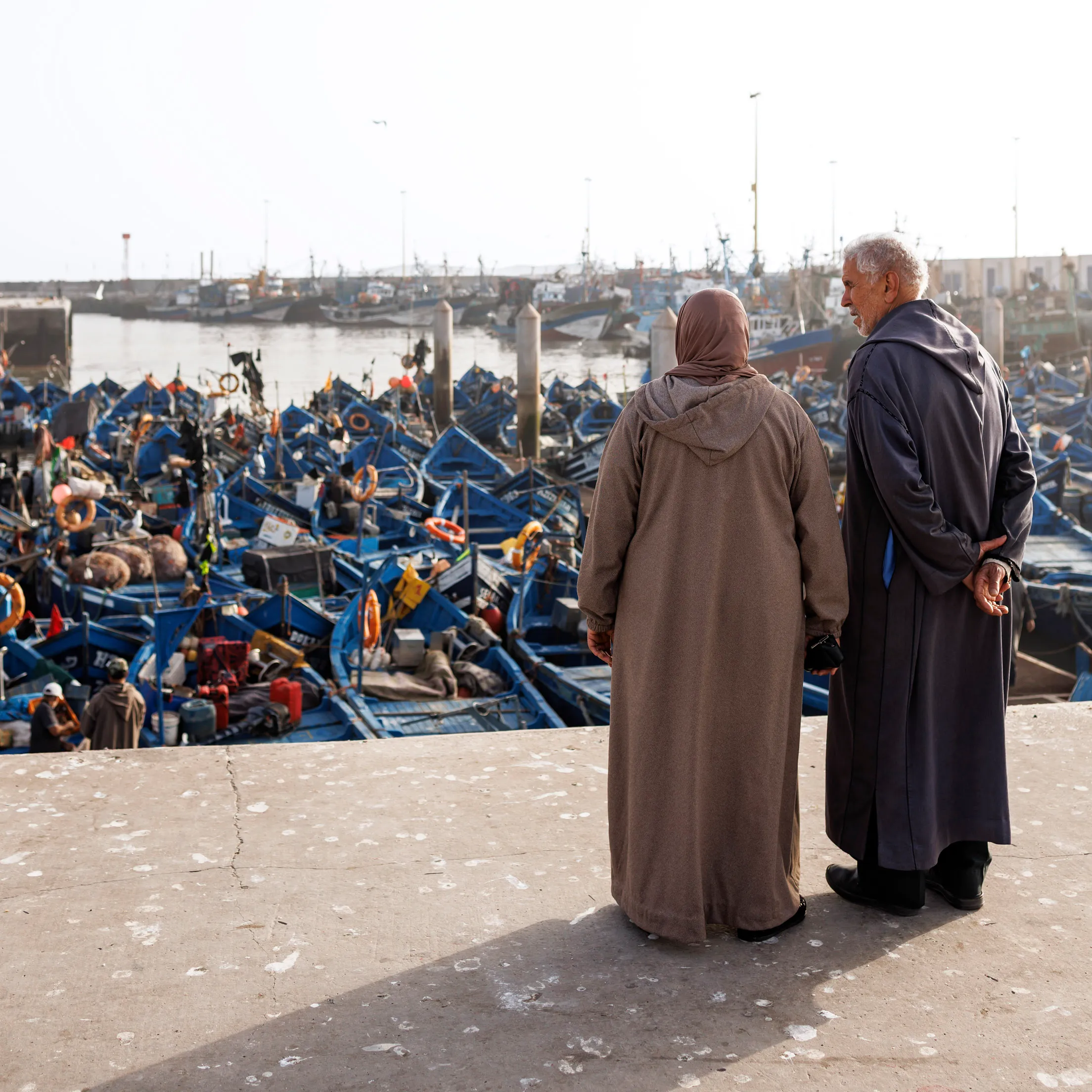 Ein marokkanischer Mann und eine Frau mit langer Kleidung, stehen mit dem Rücken zum Betrachter in dem Hafen von Essaouira in Marokko. Sie schauen auf die blauen Fischerboote. Die Sonne scheint von der Seite.