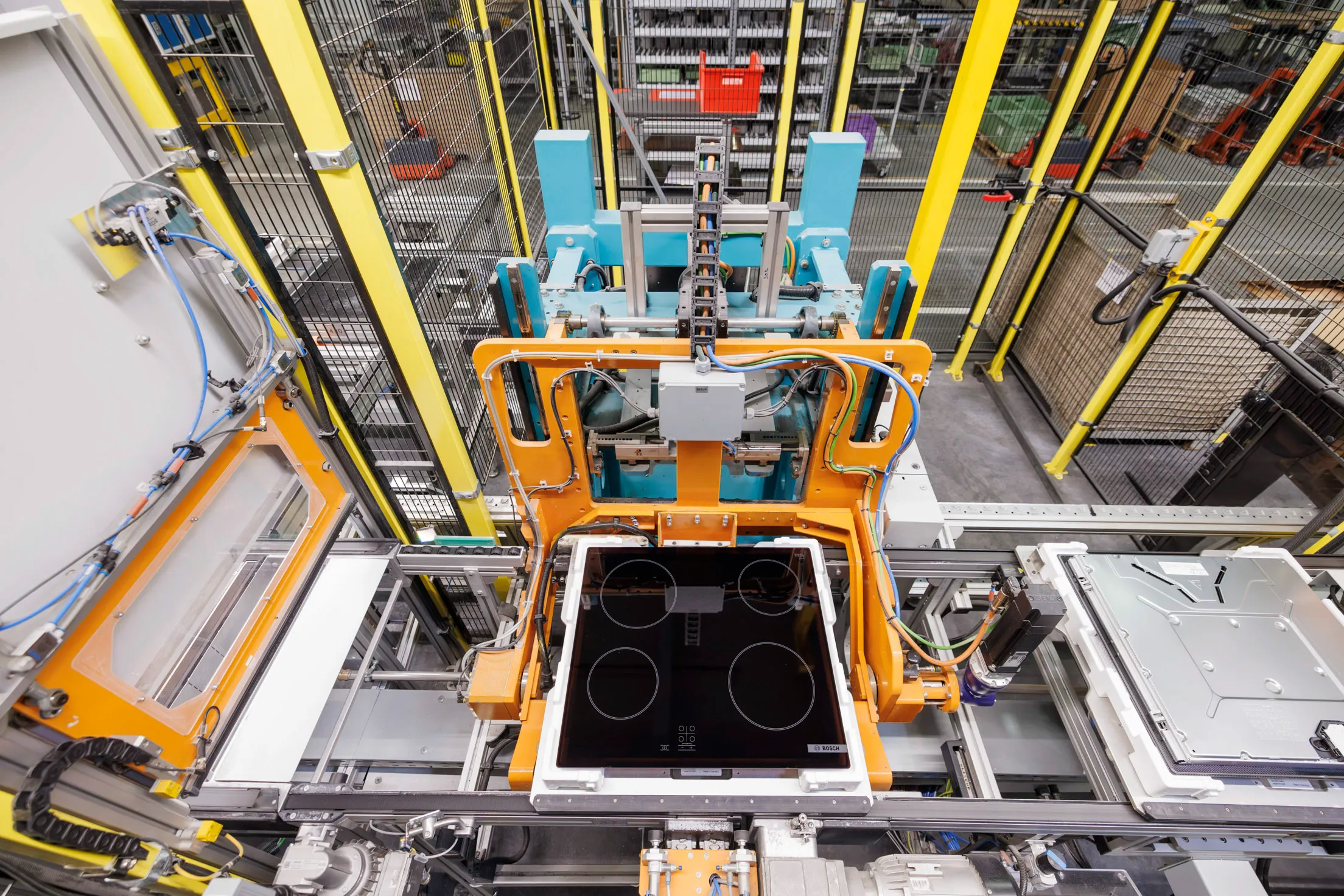 Es ist ein Fließband mit verschiedenen Cerankochfeldern zu sehen. Das Kochfeld wird von einem Roboterarm gehalten, im Hintergrund Industriehalle.