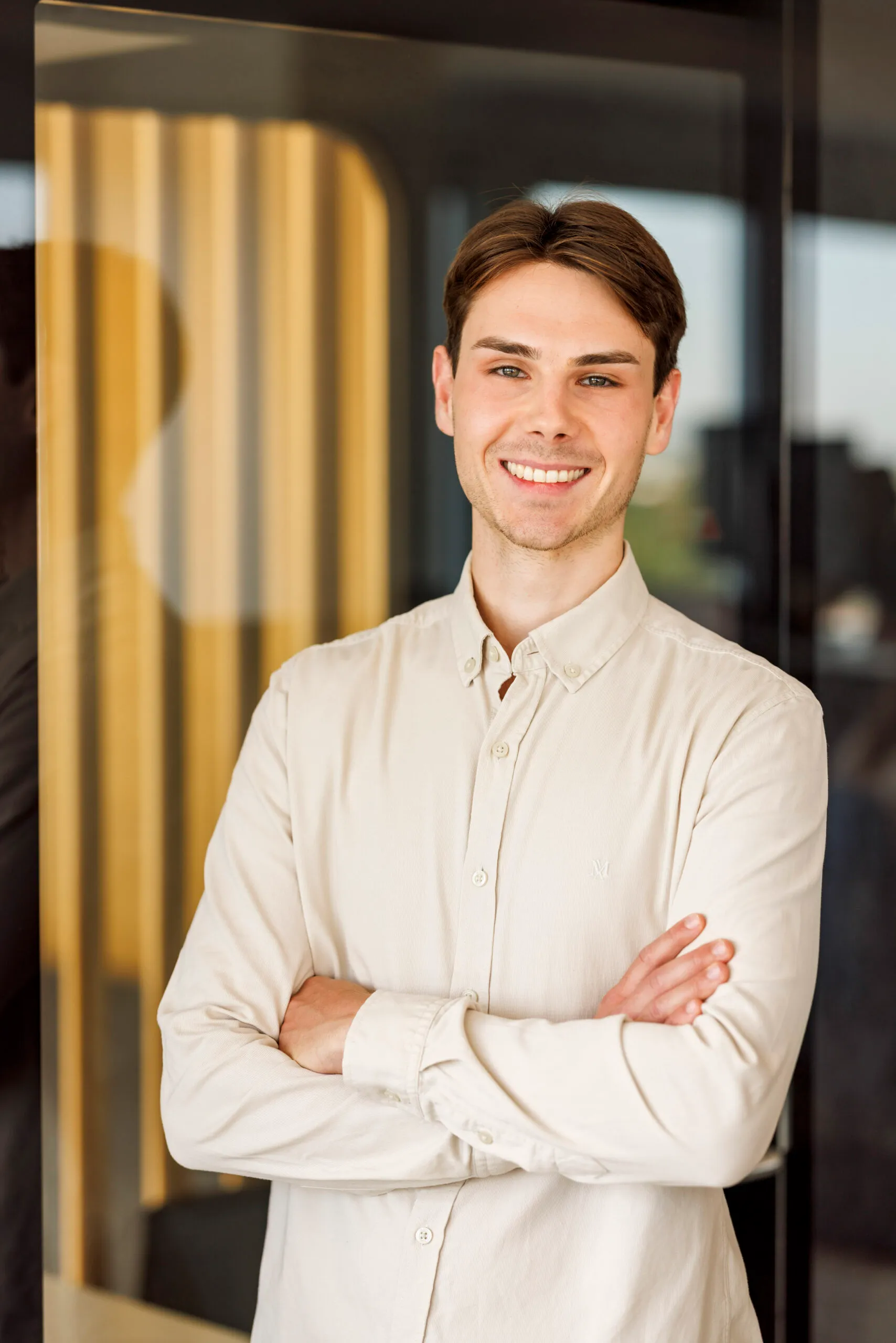 Ein junger Mann steht mit verschränkten Armen an einer Glaswand mit interessanter Spiegelung. Er hat braunes kurzes Haar und trägt ein beiges Hemd