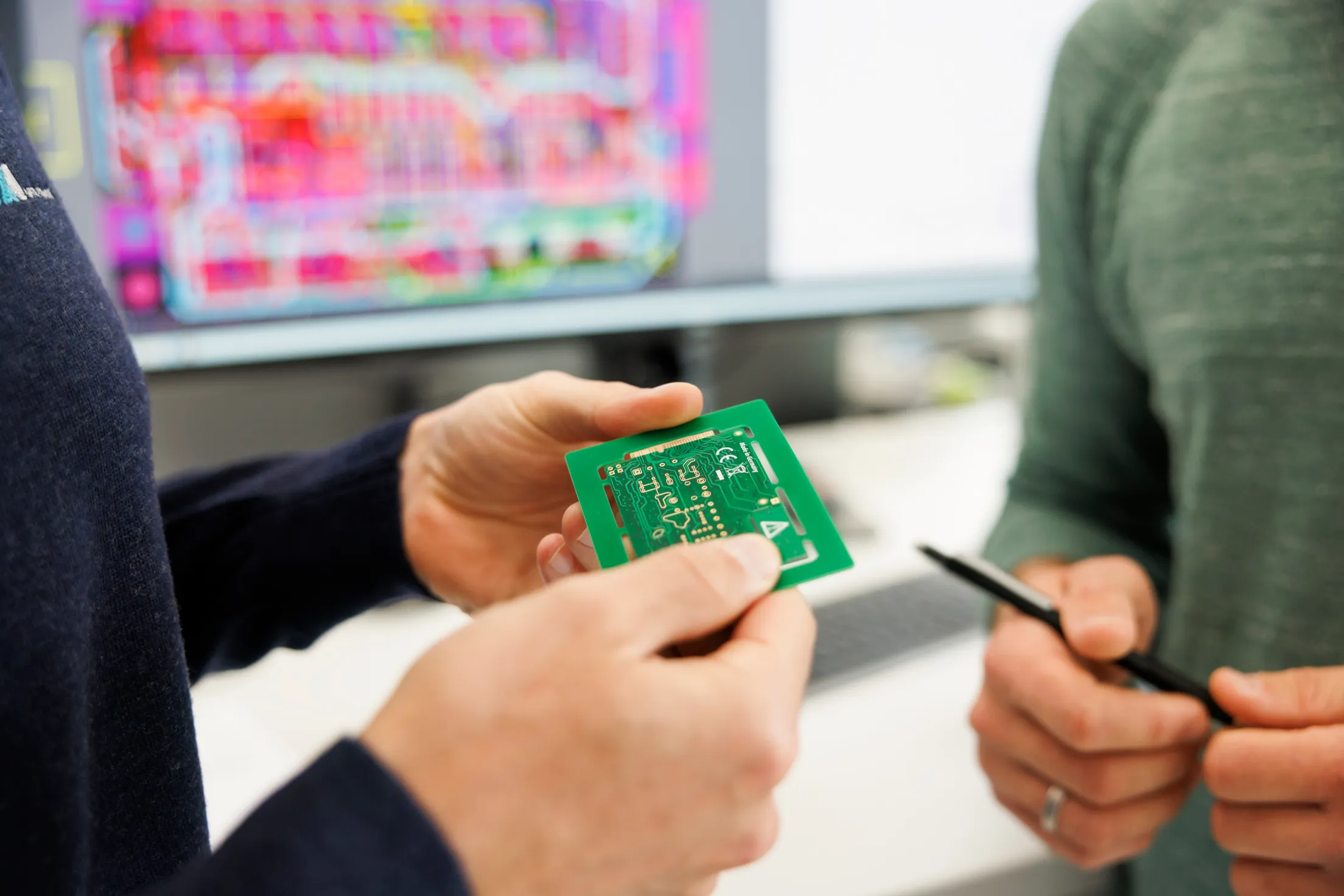 Eine Hand hält ein grünen Mikrochip mit beiden Händen. Gegenüber sieht man eine andere Hand, die einen schwarzen Stift hält.