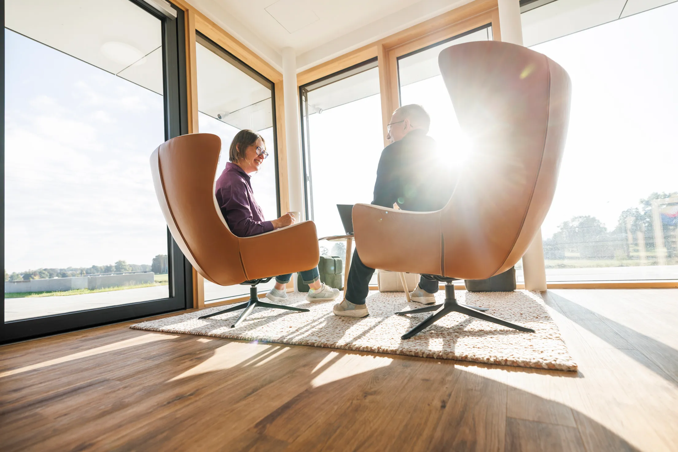 Zwei Personen sind von hinten im Gegenlicht zu sehen und sie sitzen in zwei großen, hellbraunen Sessel und unterhalten sich. Das Foto ist leicht von unten aufgenommen und durch die Fensterfront strahlt die Sonne schön ins Bild.