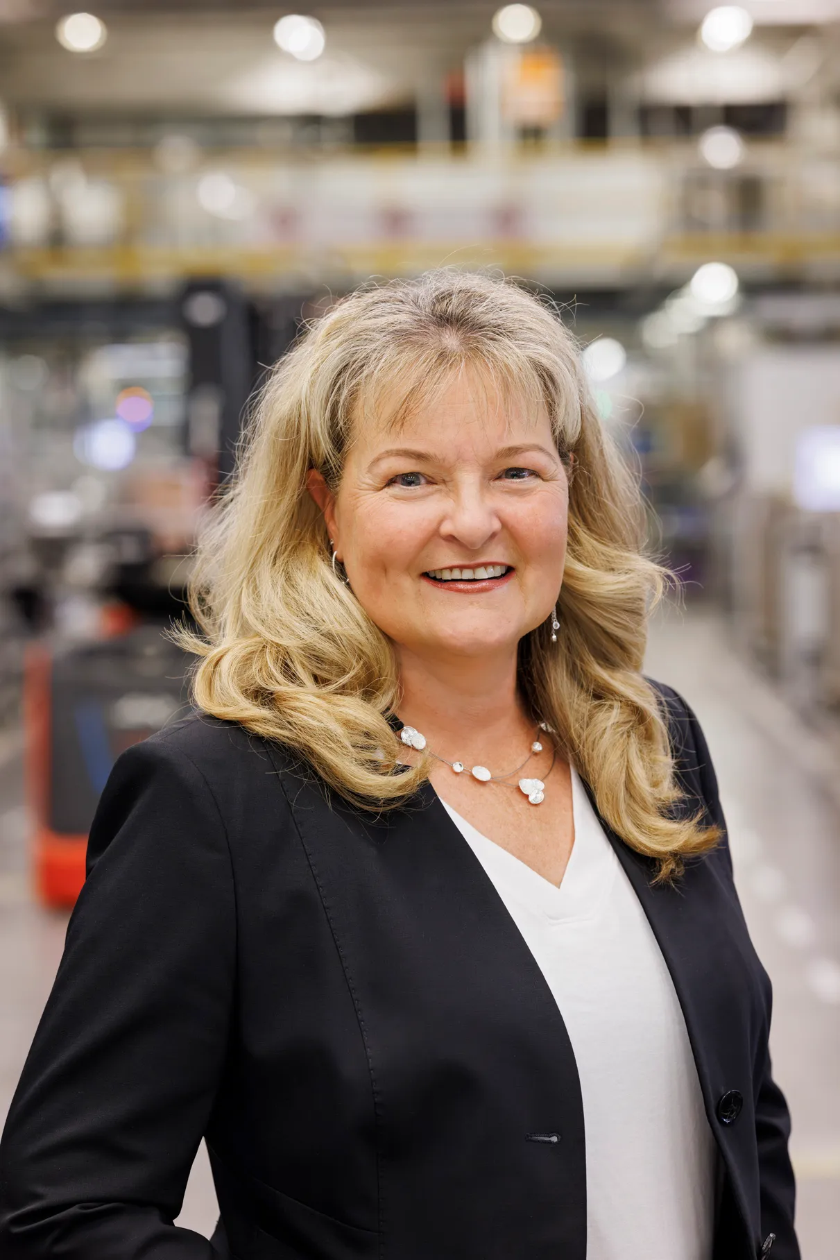 Eine Frau mit längerem, blonden Haar steht in einer Fabrikhalle. Sie trägt ein schwarze Jacket und ein weißes Shirt, im Hintergrund ist unscharf eine Industriestrasse zu sehen.
