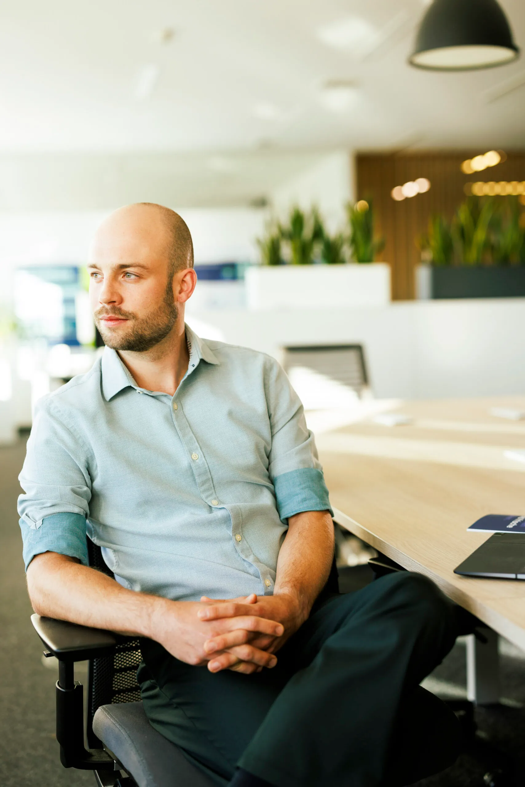Ein junger Mann mit einem türkisfarbenen Hemd und ganz kurzem Haar und Bart, sitzt an einnem Konferenztisch auf einem Stuhl und schaut aus dem Fenster