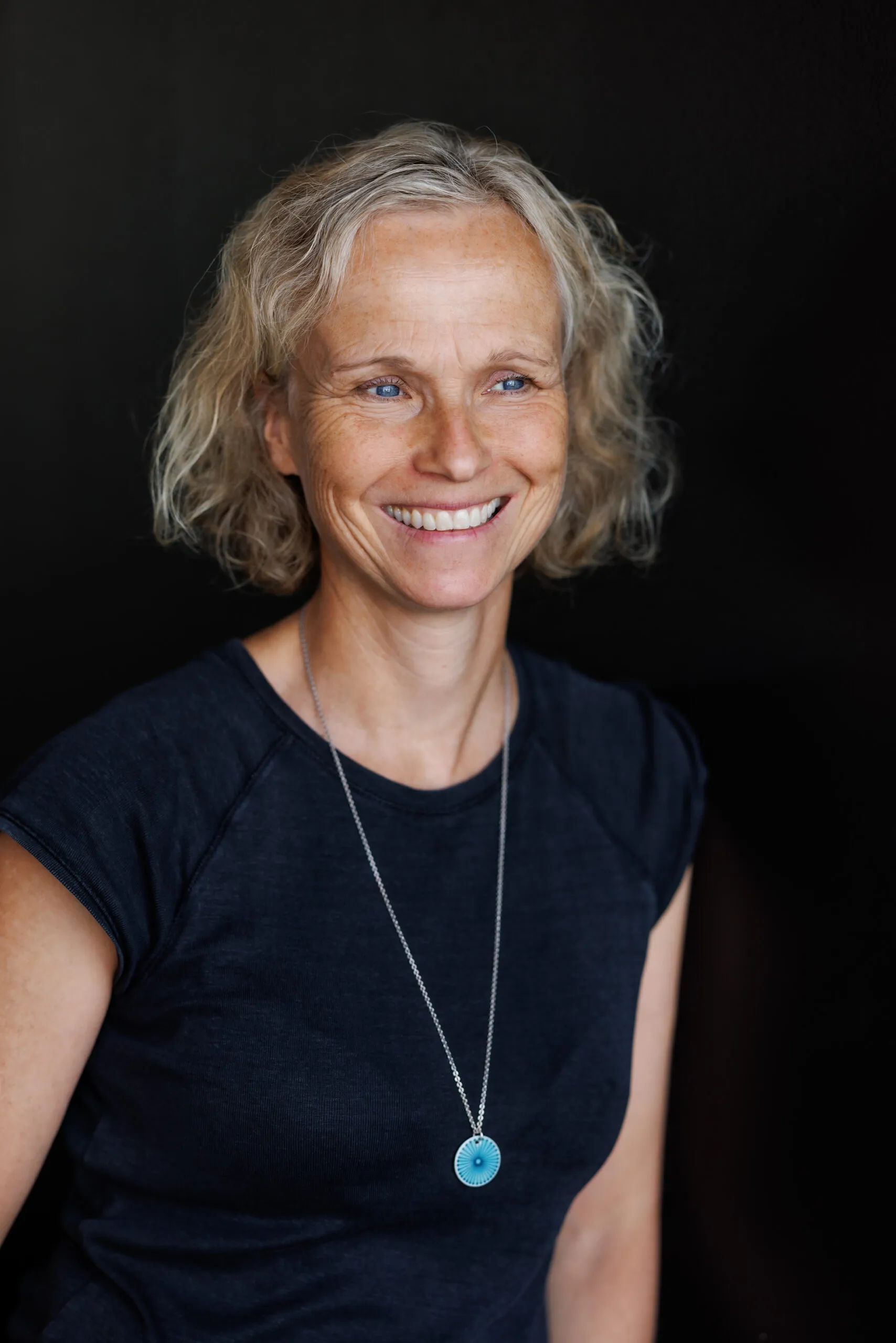 Ein Portraits der Fotografin Carolin Jacklin, sie ist mit dem Oberkörper abgebildet und schaut lachend seitlich nach rechts. Sie hat blondes, kinnlanges Haar, trägt ein dunkelblaues T-Shirt und eine lange, silberne Kette mit einem hellblauen Anhänger. Der Hintergrund ist schwarz.