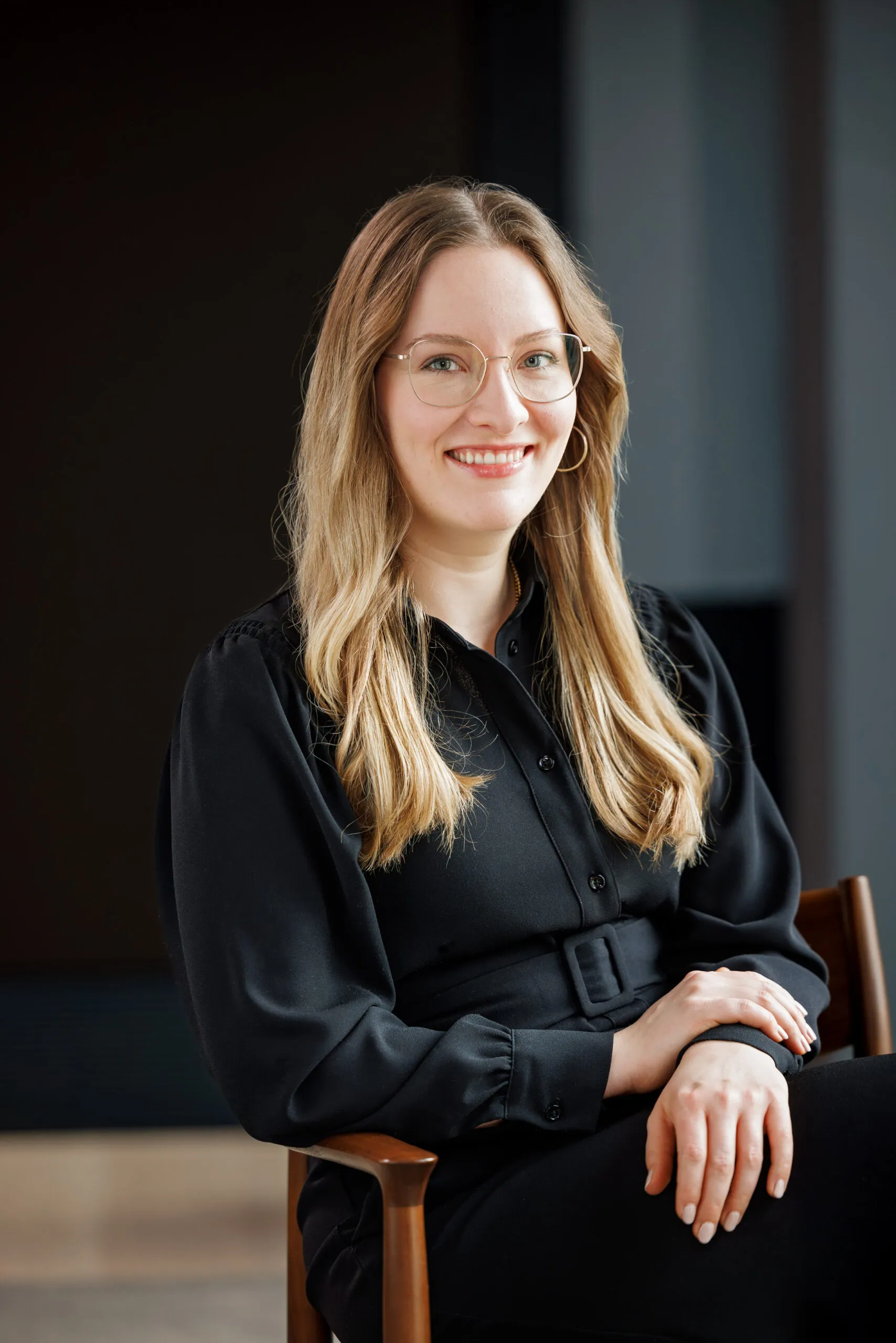 Portraits einer jüngeren Dame, die auf einem Stuhl sitzt und in die Kamera lächelt. Sie hat dunkelblondes, langes Haar, eine goldene Brille und ein schwarzes Kleid an. Hintergrund ist schwarz und grau.