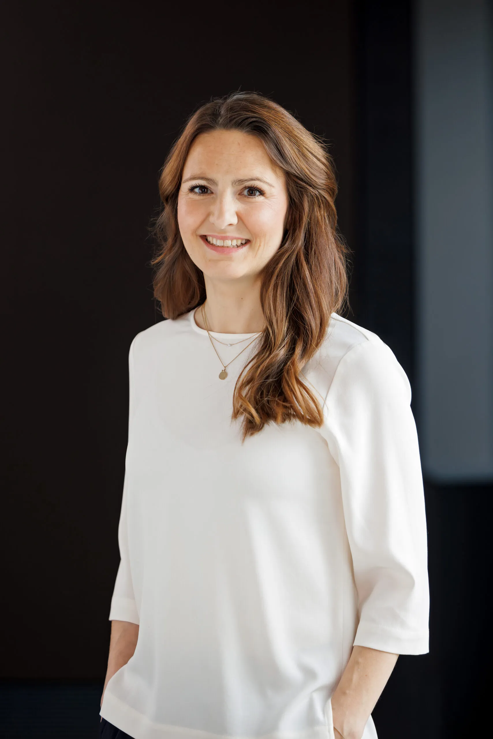 Portraits einer jüngeren Dame mit braunem, langem Haar. Sie trägt ein weißes Shirt, Goldkette mit Anhänger und lächelt in die Kamera. Hintergrund ist schwarz und grau.