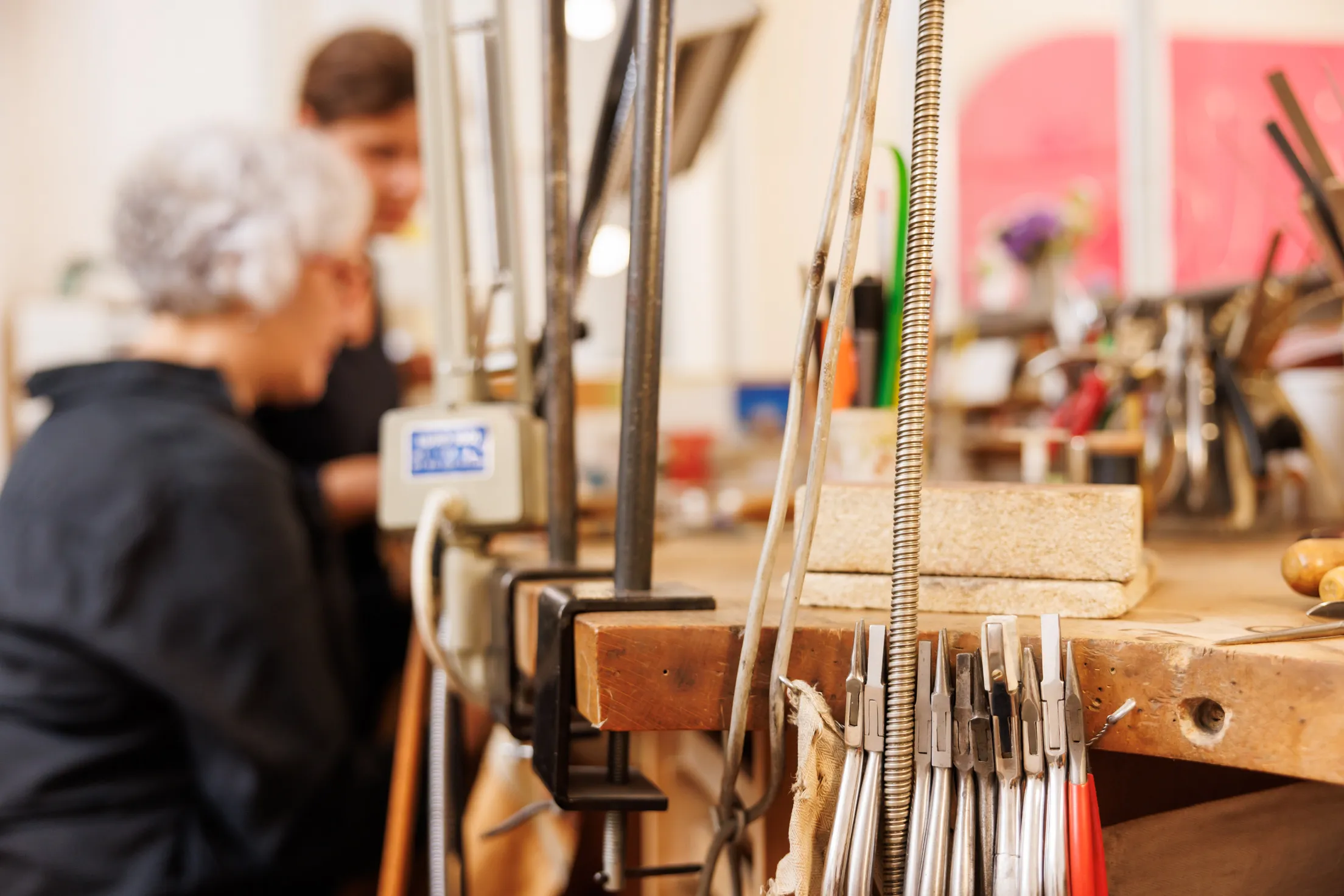 In einer Werkstatt liegt die Schärfe auf Werkzeug, viele Zangen und ein Arbeitstisch ist zu sehen. Im Hintergrund unscharf zwei Goldschmiedinnen in dunkler Kleidung bei der Arbeit, eine Dame sitzt, die andere Dame steht dahinter.
