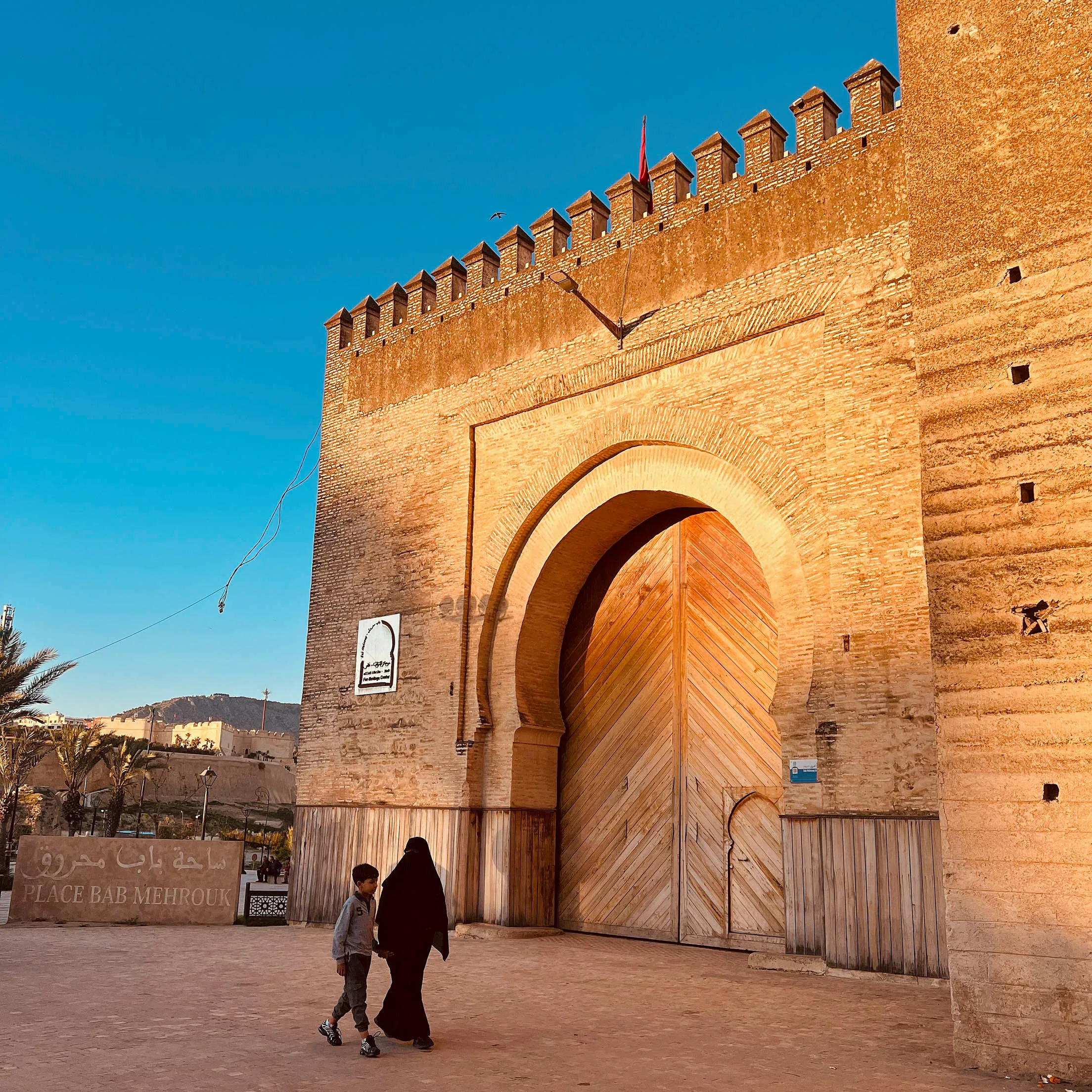 Zwei Personen, eine Frau mit schwarzer Burka und ein Junge, gehen auf einem Platz spazieren. Dahinter in goldenem Licht angestrahlt, befindet sich eine Mauer mit einem großen Tor. Es ist blauer Himmel zu sehen.