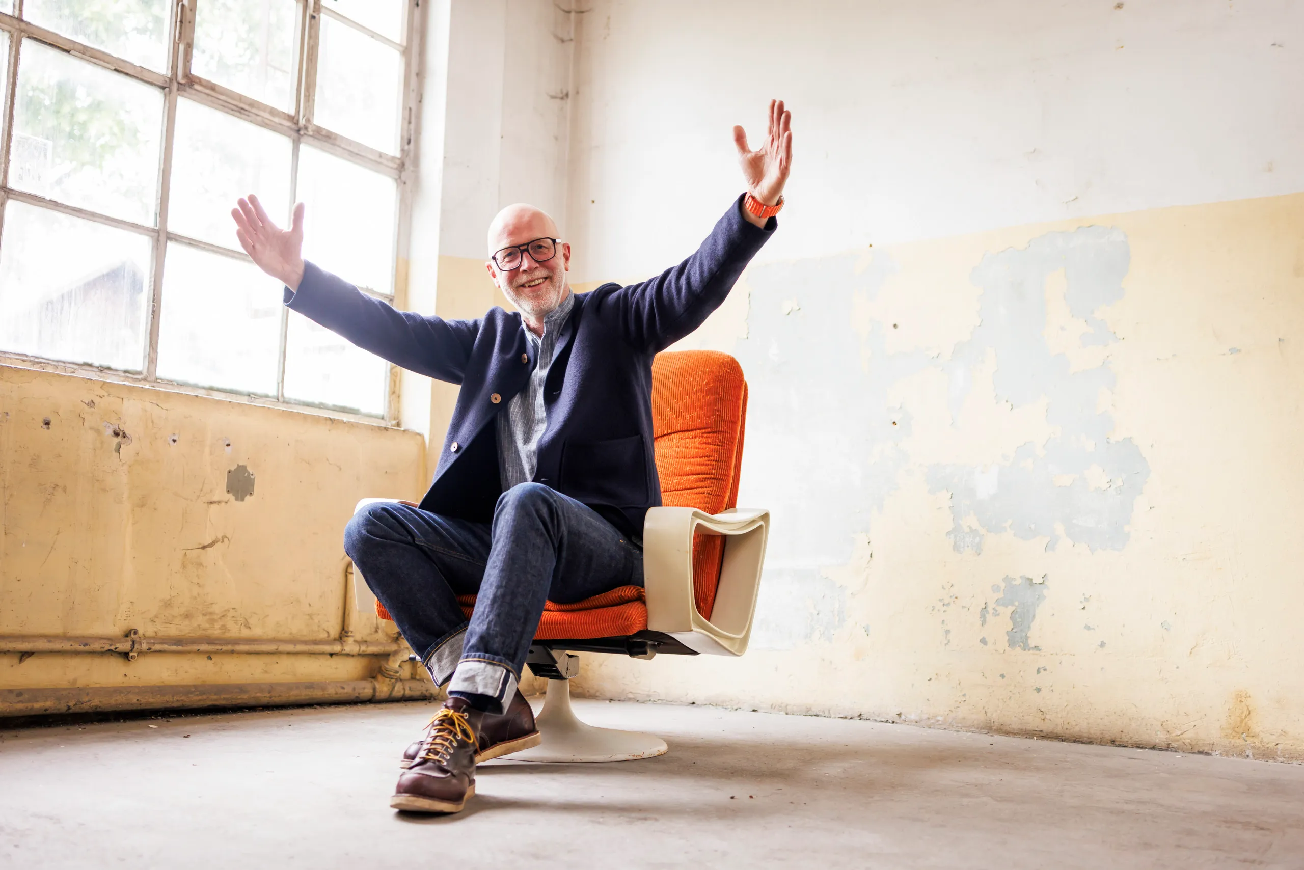 Mann mit grau meliertem Bart und dunkler, großer Brille sitzt in einem orangenen Sessel und er hebt lachend die Arme hoch. Er wirkt sehr freundlich und einladend in einem leeren, runtergekommenen Raum