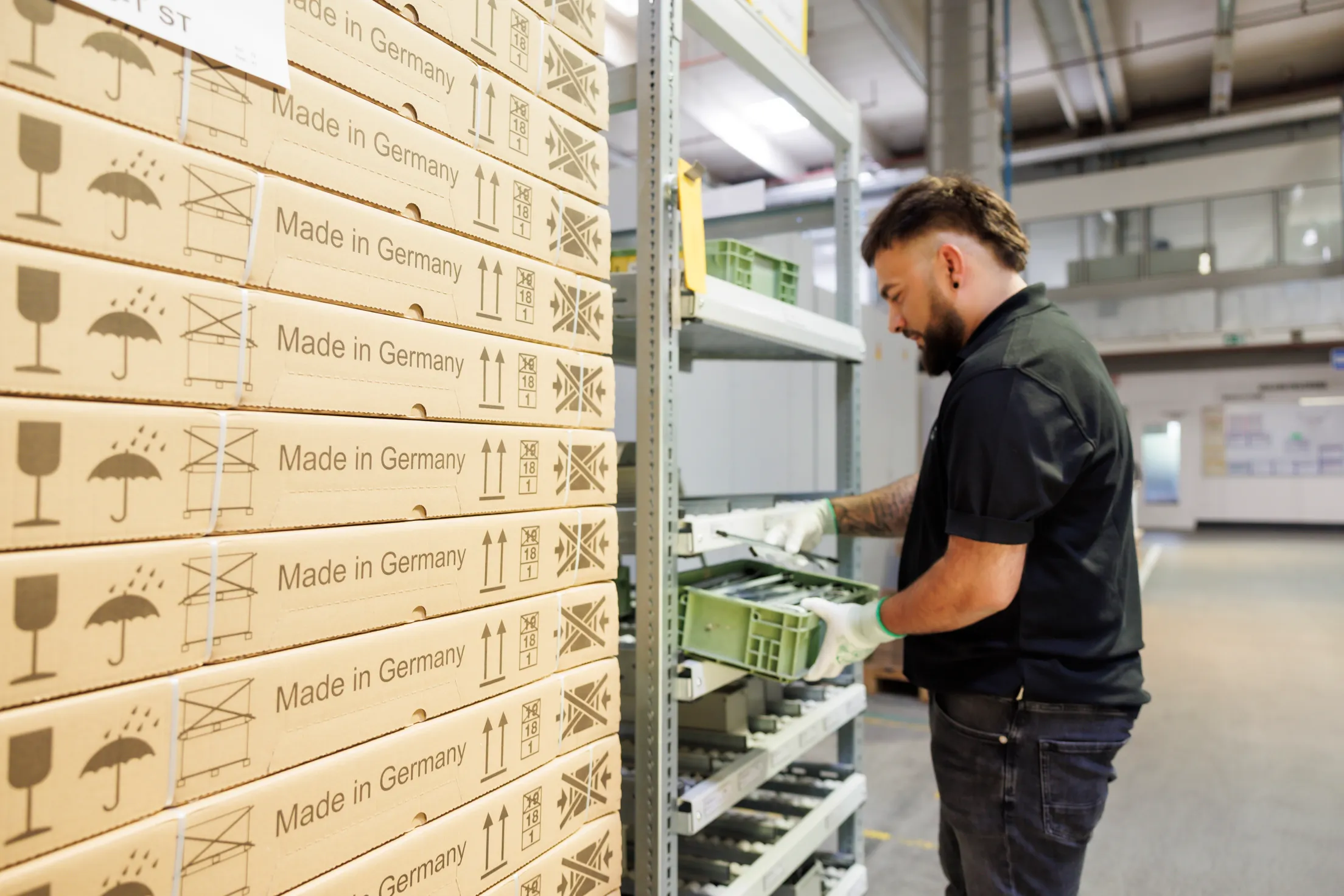 Ein Fabrikmitarbeiter schwarz angezogen, steht an einem Regal, ist unscharf und er zieht eine grüne Schublade aus dem Regal. Vorne links sind scharf Pappkartons zu sehen.