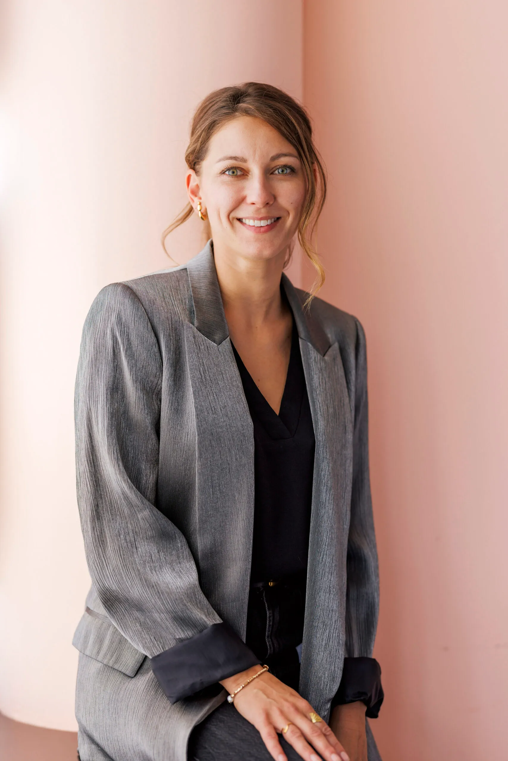 Eine junge Frau lehnt lässig an einer rosa Wand und lächelt in die Kamera. Sie trägt ihre Haare zusammengebunden, hat ein graues Jacket und ein schwarzes Shirt an.