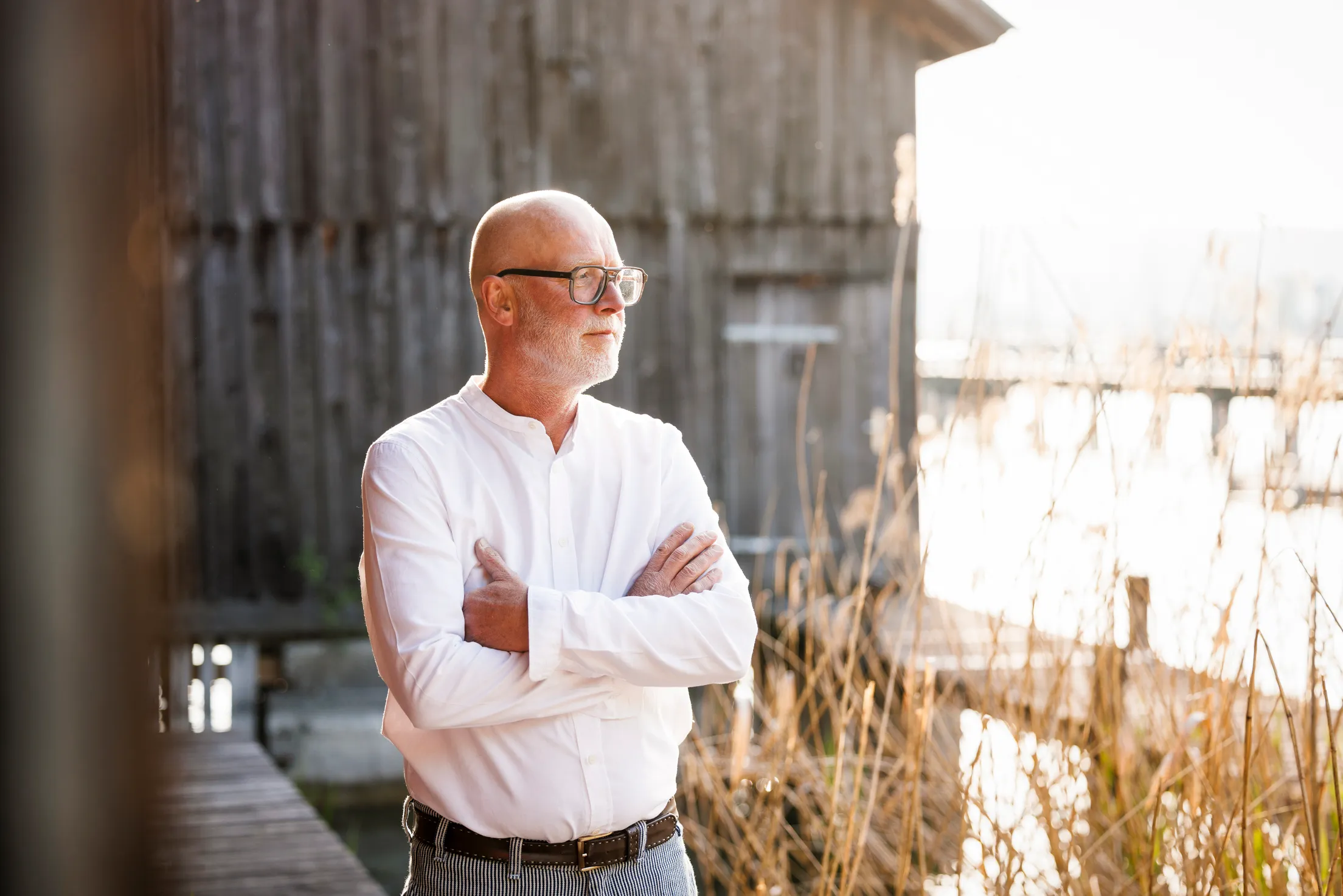 Mann mit Glatze, Brille und Bart steht mit verschenkten Armen vor einem Bootshaus am See. Er trägt ein weißes Hemd und schaut nacht rechts im Bild