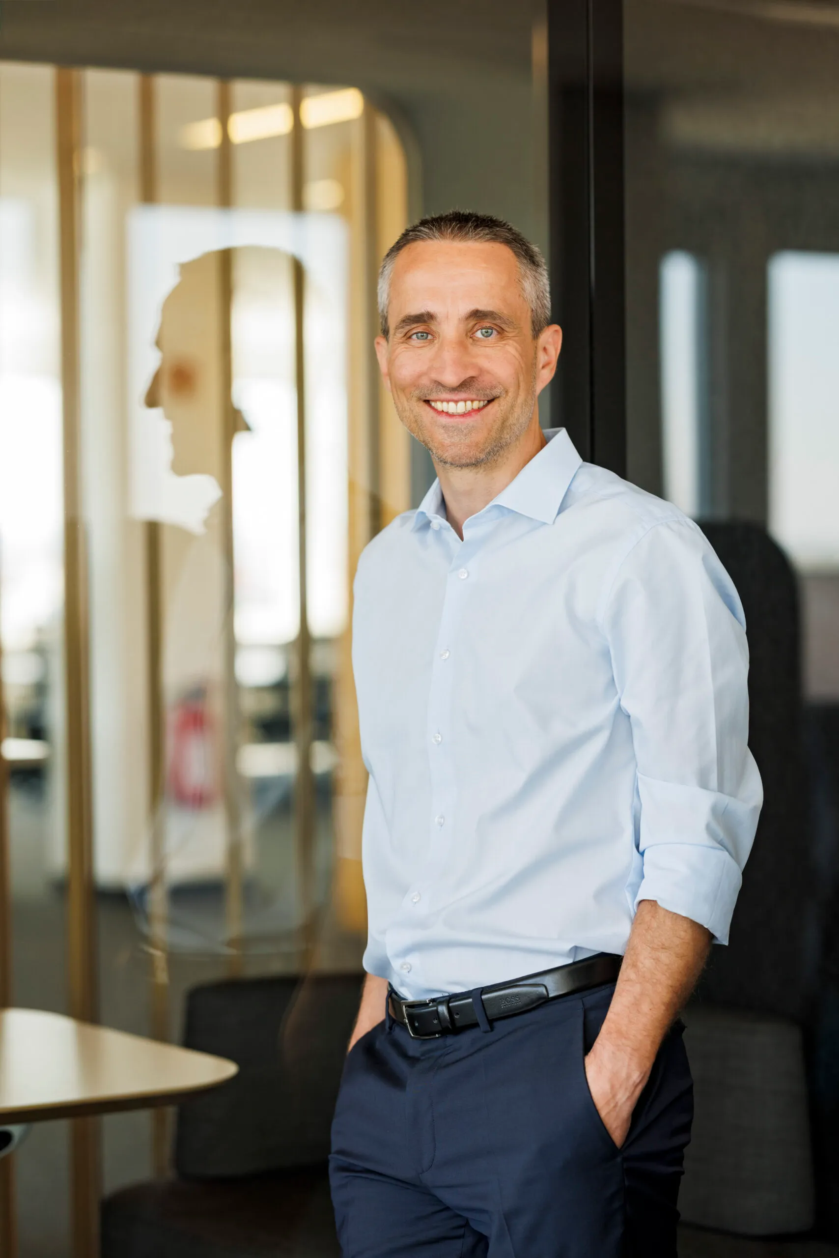 Ein Mann mit schwarz grauem, kurzem Haar, steht mit einem hellblauen Hemd an eine Glastür gelehnt mit einer interessanten Spiegelung im Glas. Er hat die Hände in den Hosentaschen und steht selbstbewusst da