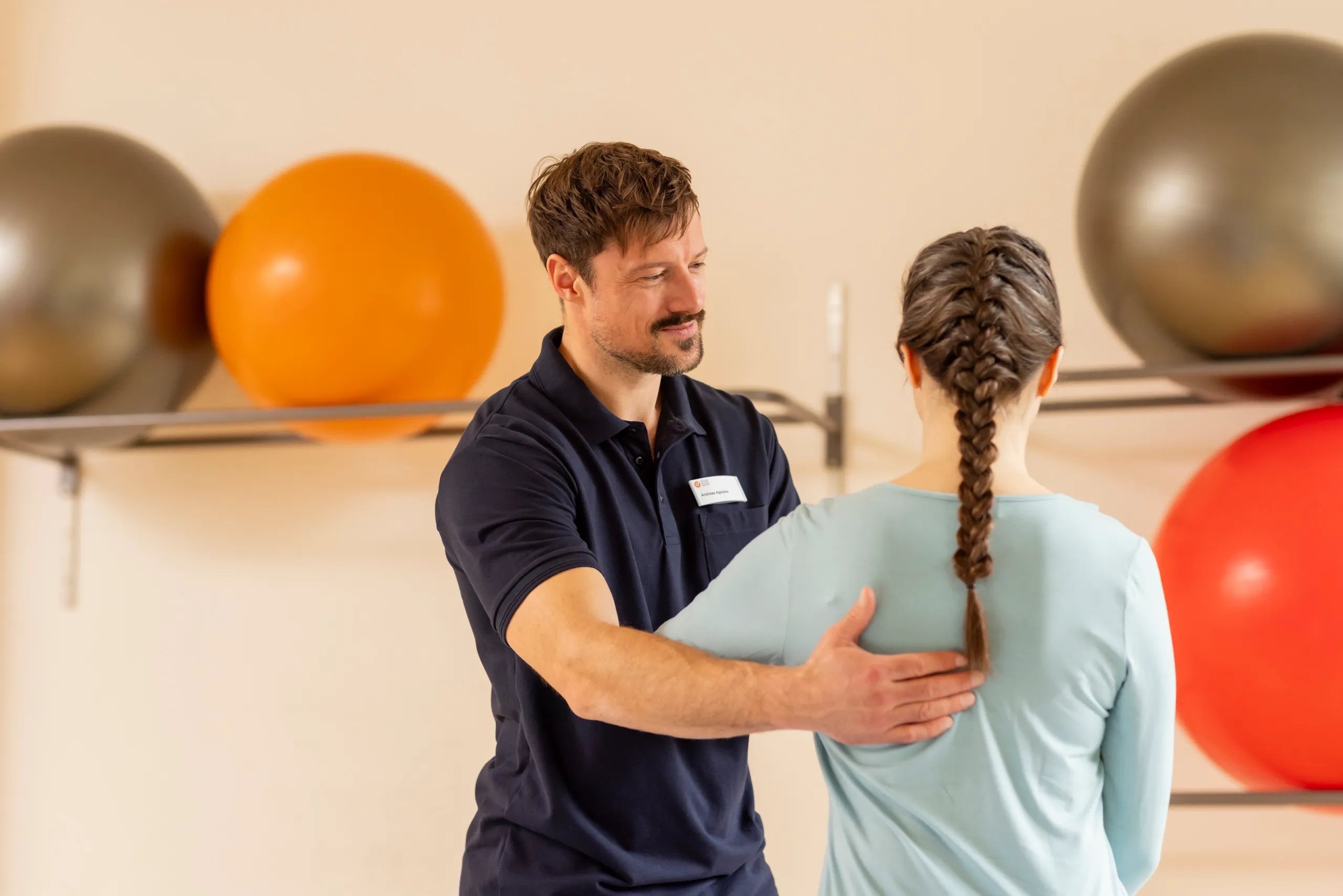 Ein Physiotherapeut mit einem dunkelblauen Poloshirt behandelt eine Dame mit türkisfarbenen Shirt an ihrer Schulter. Sie trägt einen geflochtenen Zopf und sitzt mit dem Rücken zur Kamera. Im Hintergrund sind Medizinbälle unscharf zu sehen.