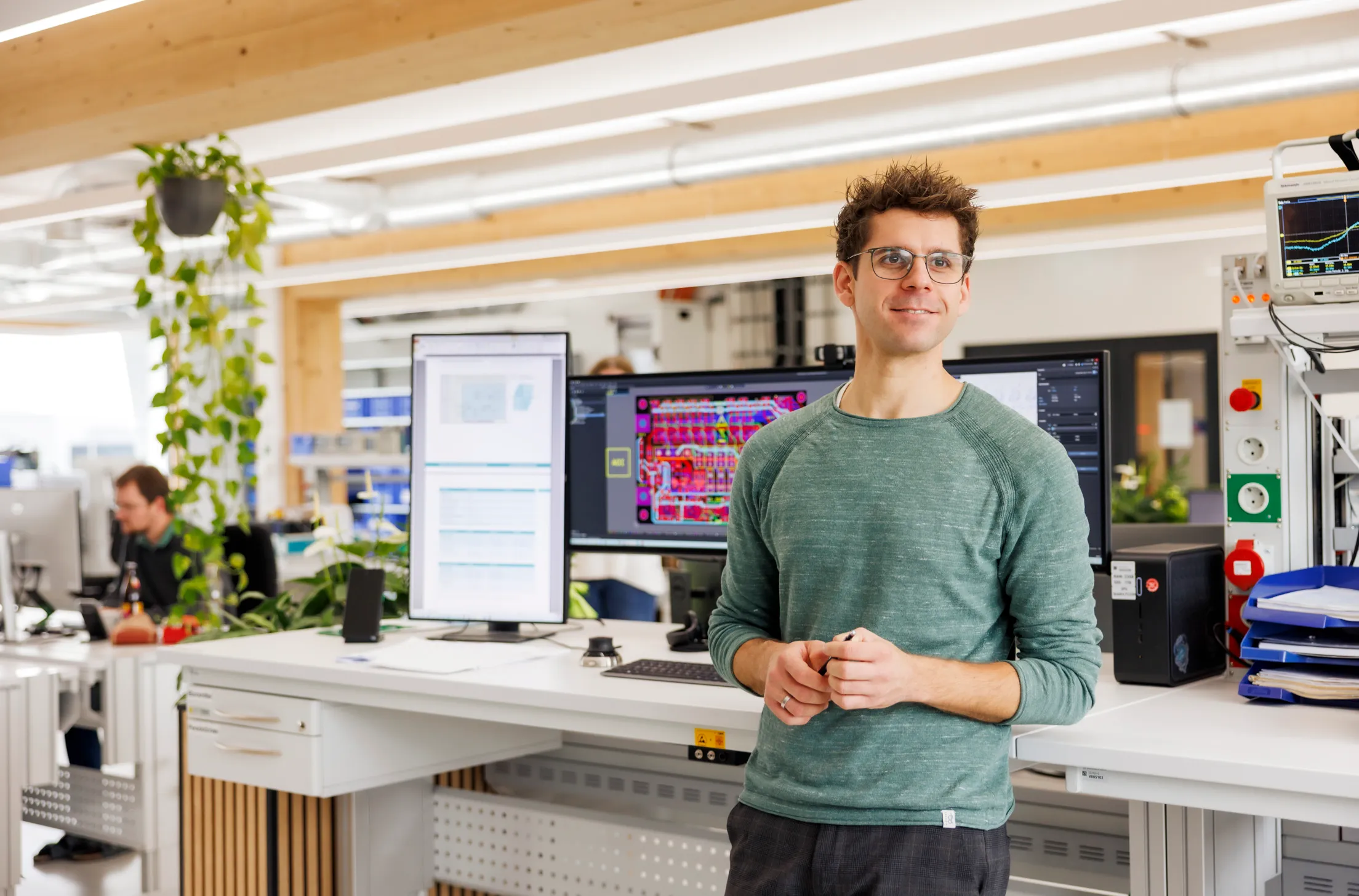 Ein sympathisch lächelnder Mann mit grünem Sweatshirt steht an einem hohen Schreibtisch und schaut nach links im Bild. Er steht vor einem Computer mit großen Bildschirmen.