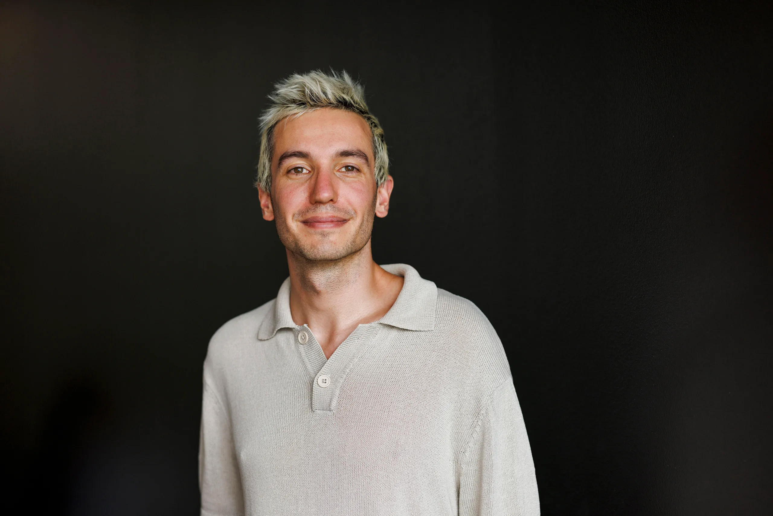 Ein jüngerer, cooler Mann schaut selbstbewusst in die Kamera und steht vor einem schwarzen Hintergrund. Er hat blond gefärbte Haare und trägt einen beigen Pullover.