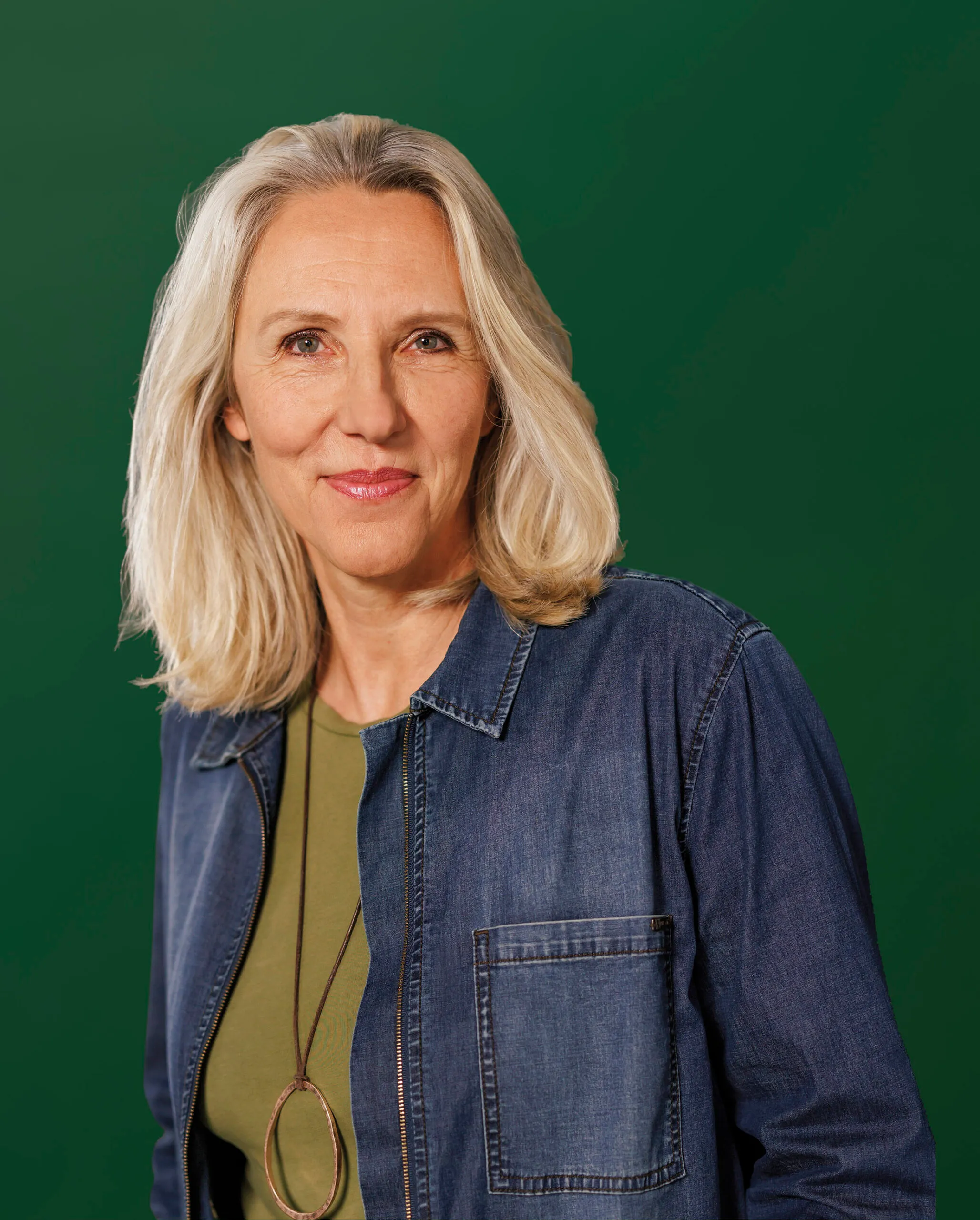 Eine Frau mit blondem längeren Haar trägt ein blaues Jeanshemd und einen grünen Pullover darunter. Sie lächelt leicht in die Kamera und steht vor einem dunkelgrünen Hintergrund.
