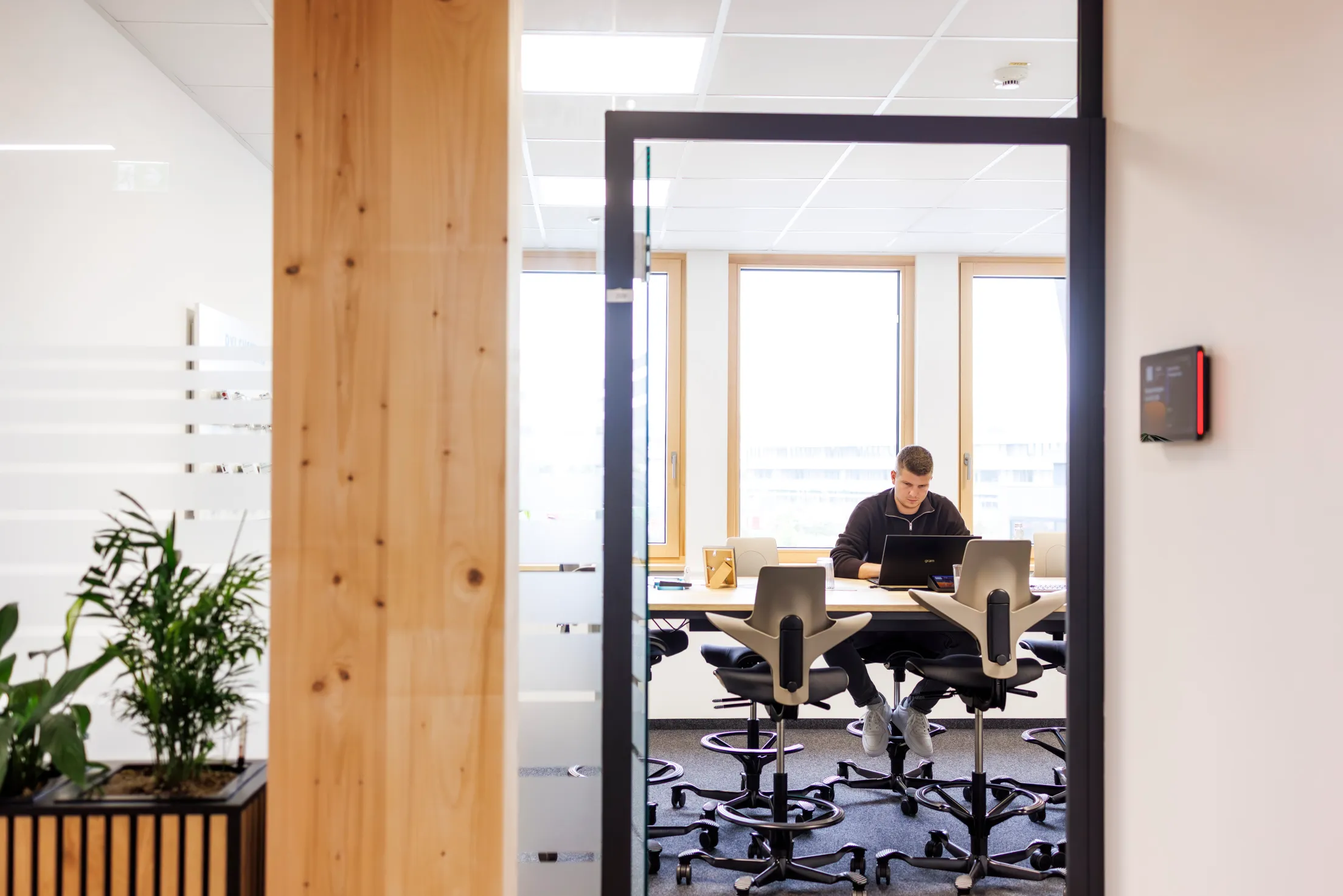 Ein Mann sitzt an einem Tisch in einem Konferenzraum und ist durch die Tür und Glasfront mit ganzem Körper auf einem Hochstuhl zu sehen. Das Foto ist sehr hell und grafisch aufgebaut.