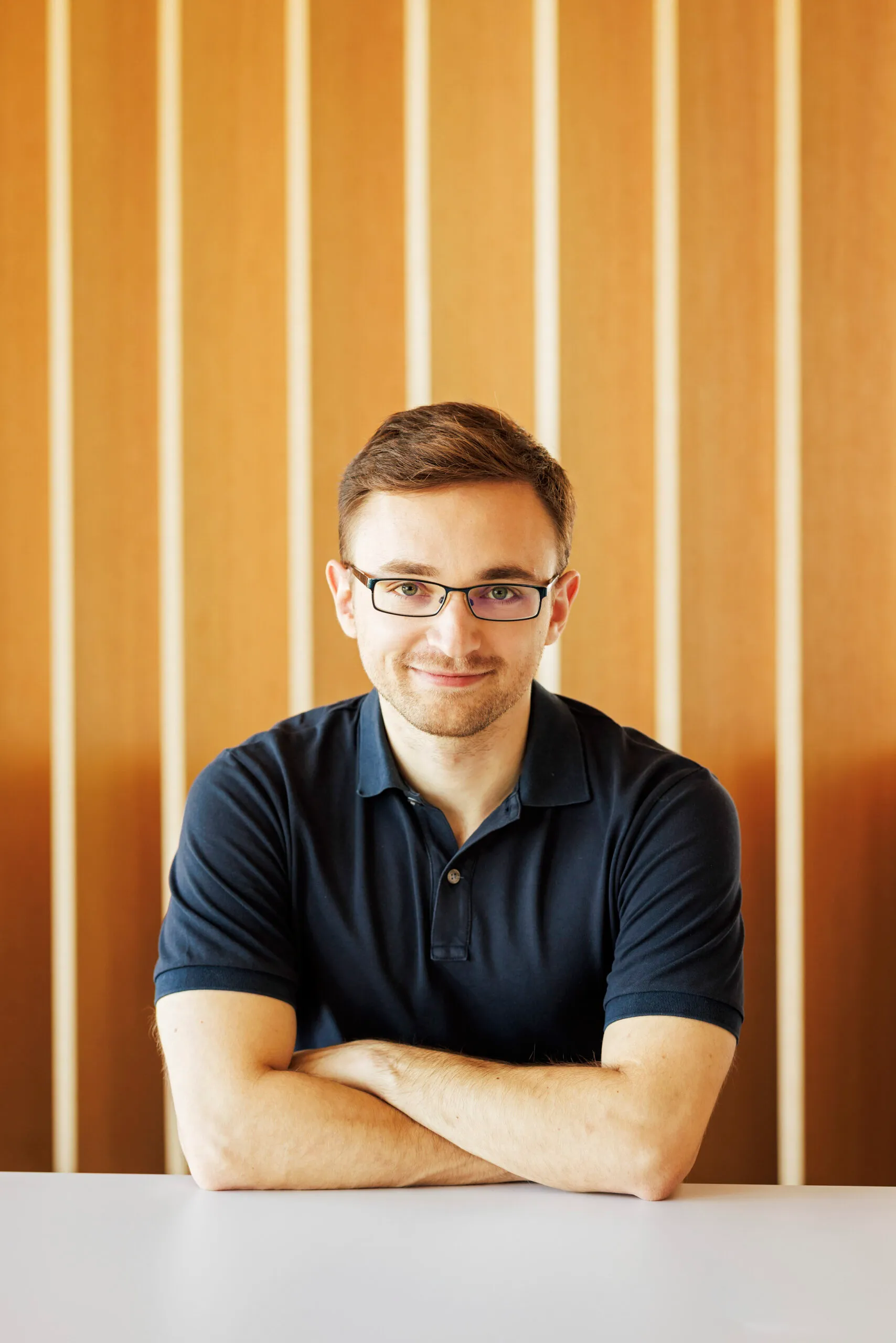 Ein junger Mann im dunkelblauen Polohemd hat die Arme verschränkt auf einem Tisch aufgestützt und schaut freundlich in die Kamera