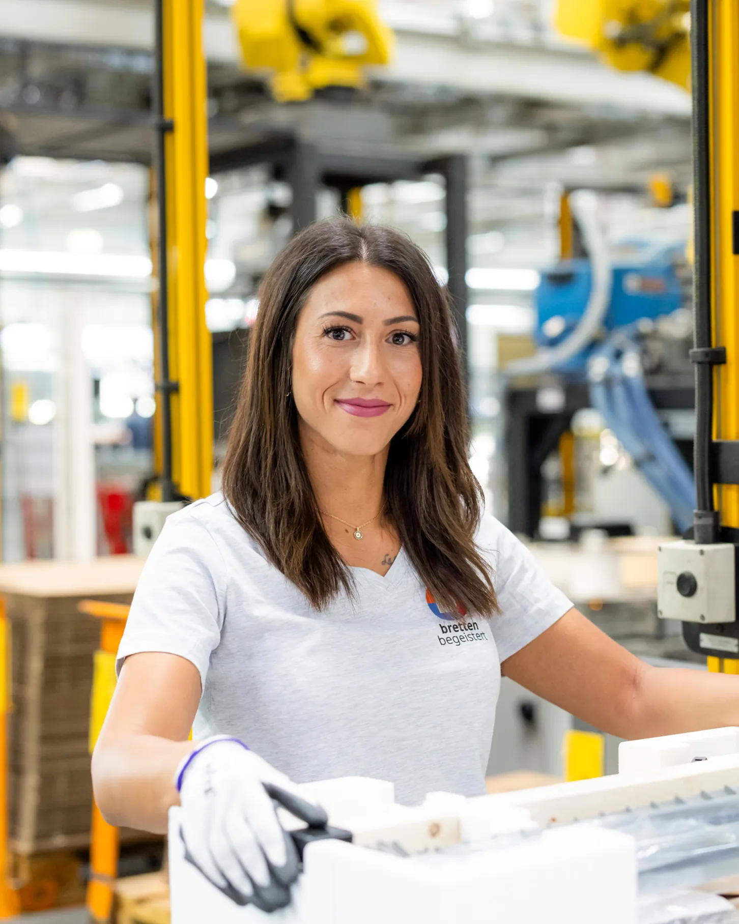 Frau mit dunklen langen Haaren trägt ein hellgraues Arbeitsshirt mit dem Logo von dem Standort Bretten. Sie steht in einer Fabrikhalle, sie hat Sicherheitshandschuhe an und packt ein fertiges Gerät in eine Styroporverpackung ein. Im Hintergrund sind unscharf Maschinen und Roboter zu erkennen.
