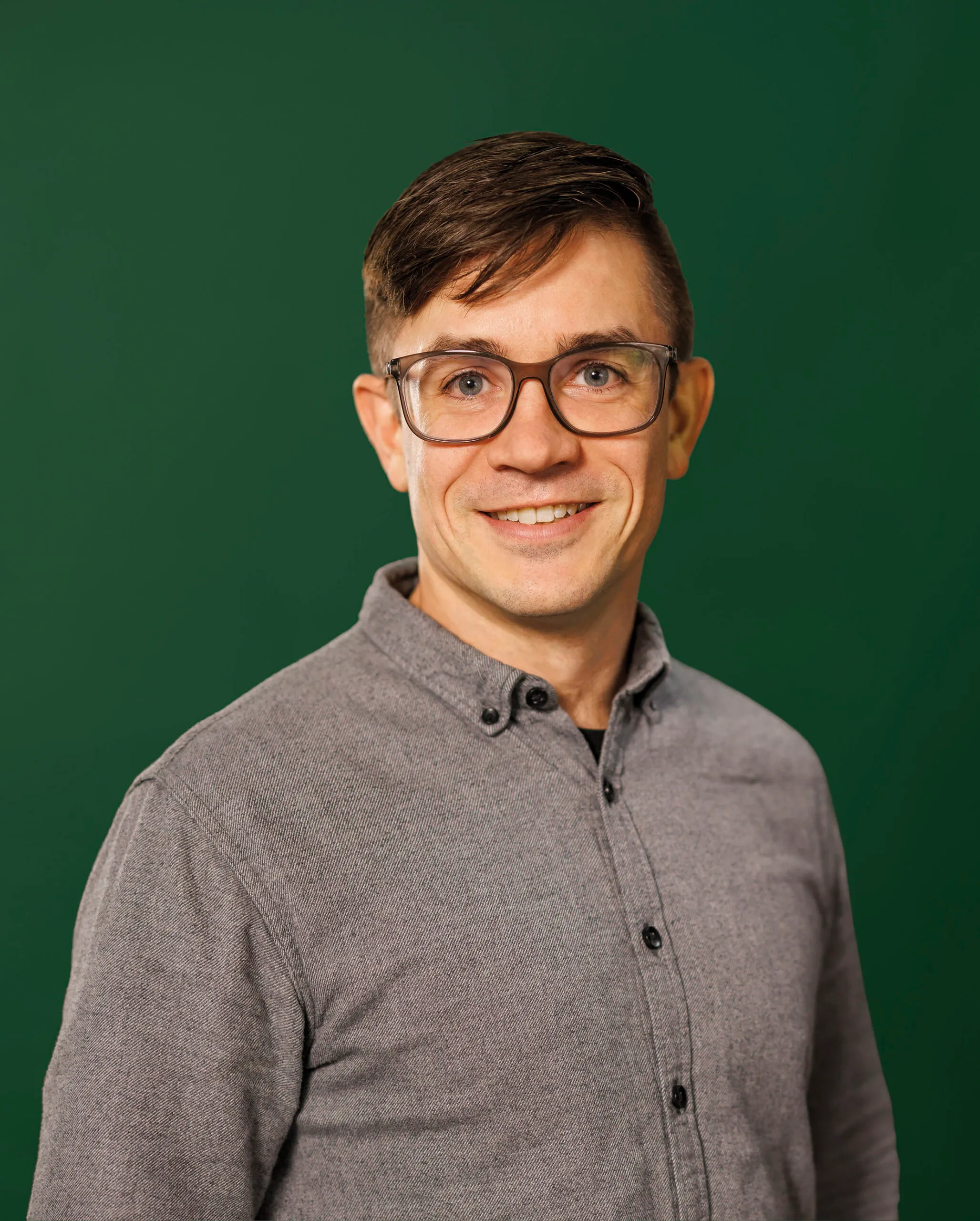 Ein jüngerer Mann mit braunem Haar und großer Brille lächelt in die Kamera. Er ist vor einem dunkelgrünen Hintergrund fotografiert und er trägt ein graues Hemd.