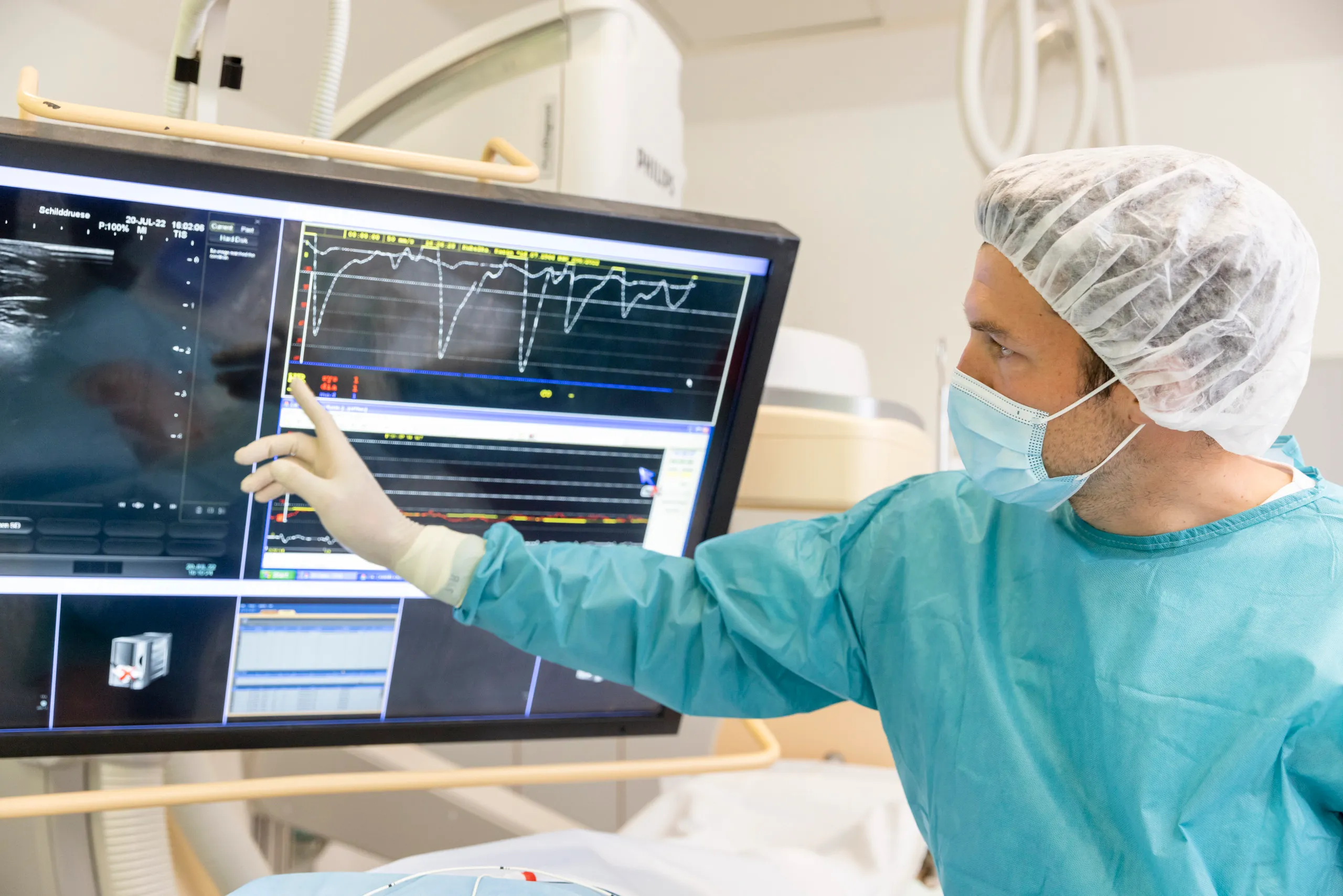 Ein Op mit einem Arzt in OP Kleidung. Er deutet auf eine Kurve auf einem Monitor, der groß im Bild zu sehen ist.