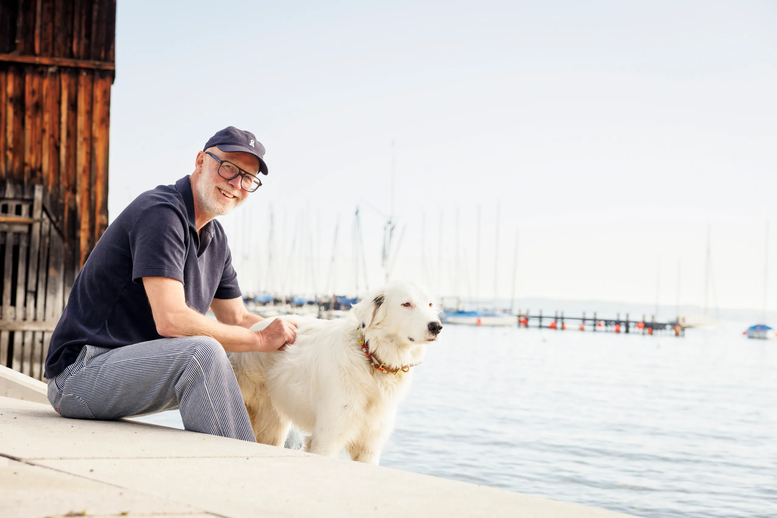 Mann mit grau meliertem Bart und großer Brille, sitzt auf einer Steintreppe an einem See. Er trägt ein blaues Cappy und blaue Kleidung und streichelt einen weißen Hund. Im Hintergrund ist ein See mit Segelbooten erkennbar.